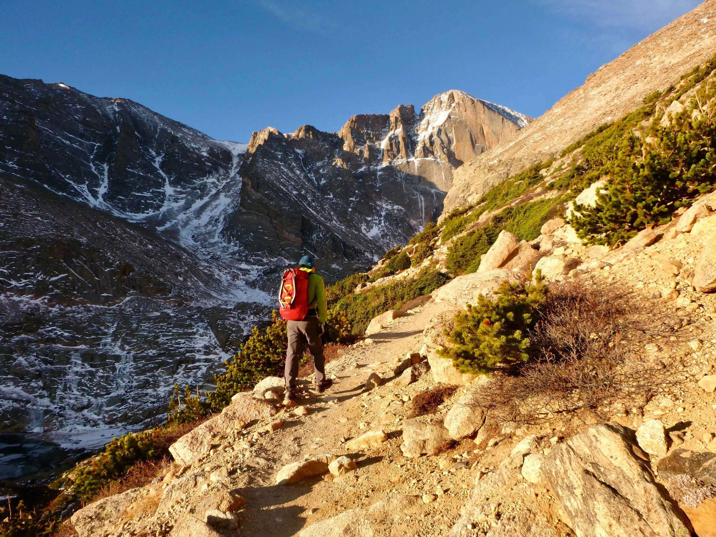 A climber wearing a red backpack with ice tools strapped to the outside is hiking the trail into Longs Peak in Rocky Mountain National Park. The icy steep face of the Diamond is in the background with early morning alpenglow.