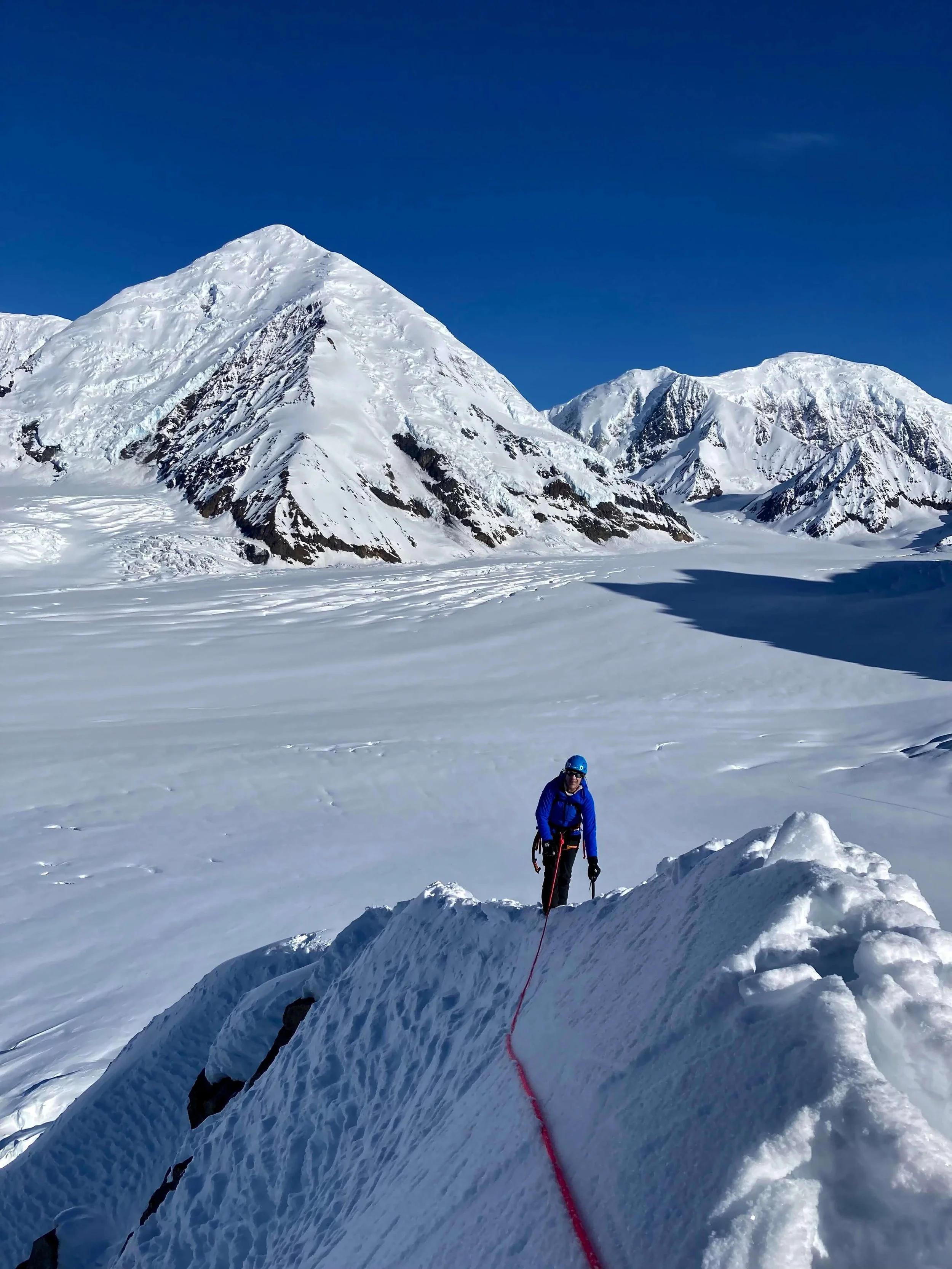 A climber in a blue jacket is traversing a knife edge snow ridge above the Kahiltna Glacier in Alaska. A snowy Mount Crosson and Kahiltna Dome are in the background.