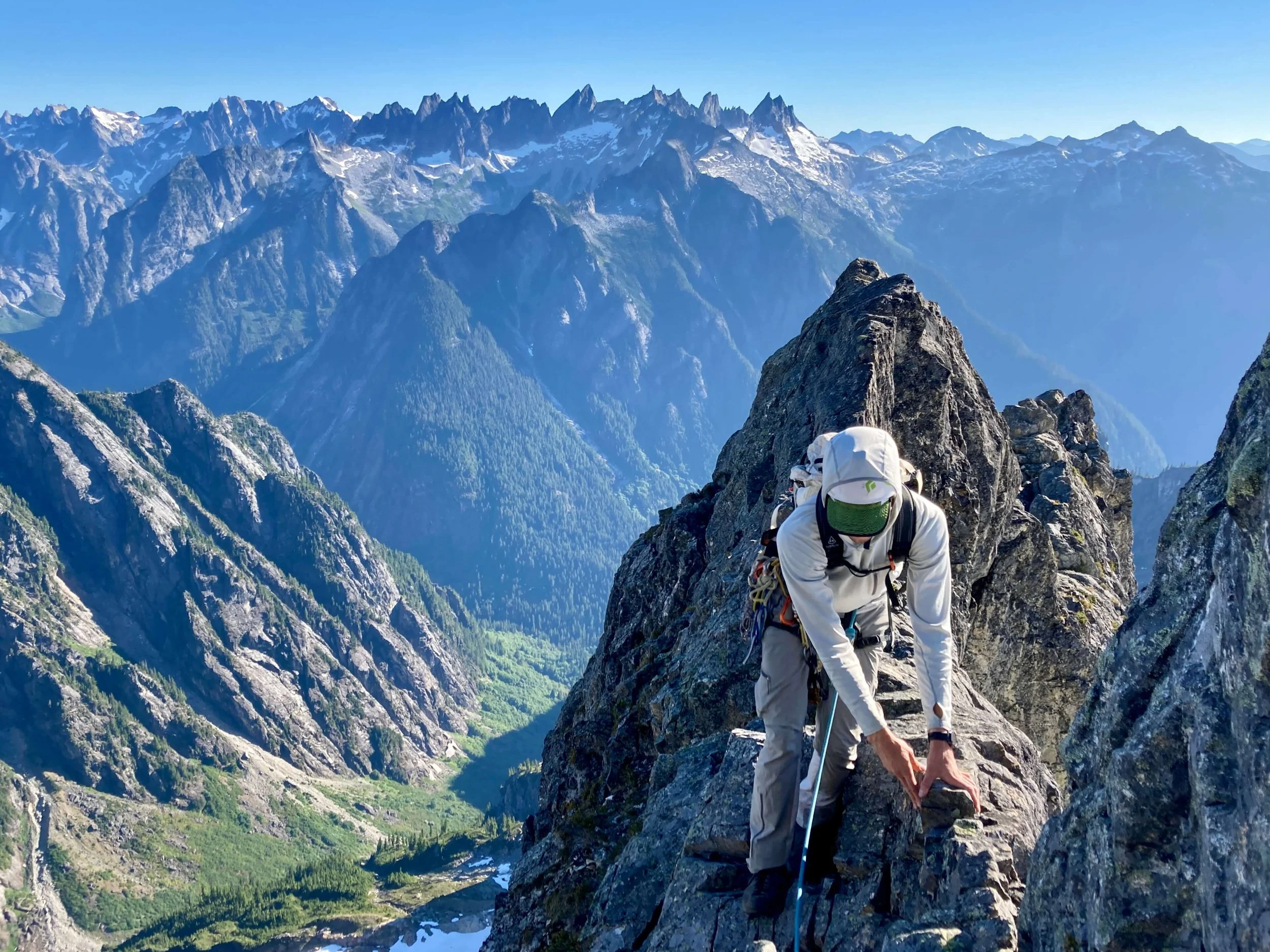 A climber in a white hooded shirt is traversing an exposed ridge on Mount Triumph with the Pickets Range in the background.