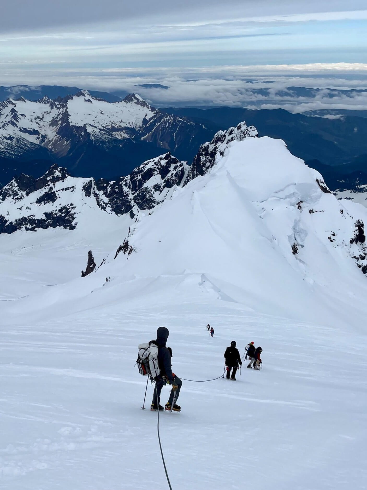 Group of climbers ascending a snowy mountain slope near a peak, with rugged, snow-covered mountain ranges and a cloudy sky in the background.