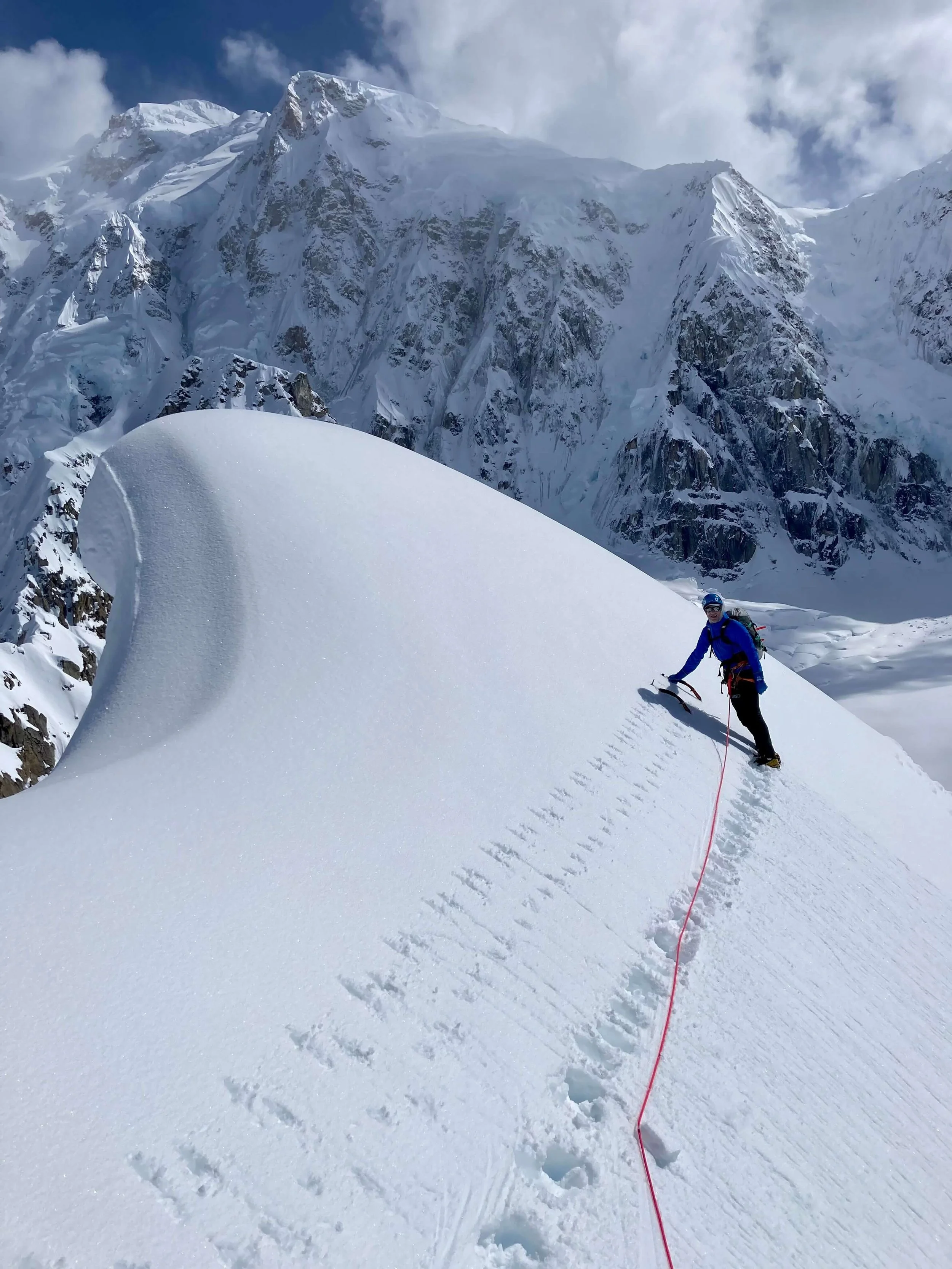 A climber in a blue jacket is traversing a steep snow slope on Annie's Ridge above the Kahiltna Glacier. The steep rocky and snowy cliffs at the base of Mount Hunter are in the background.