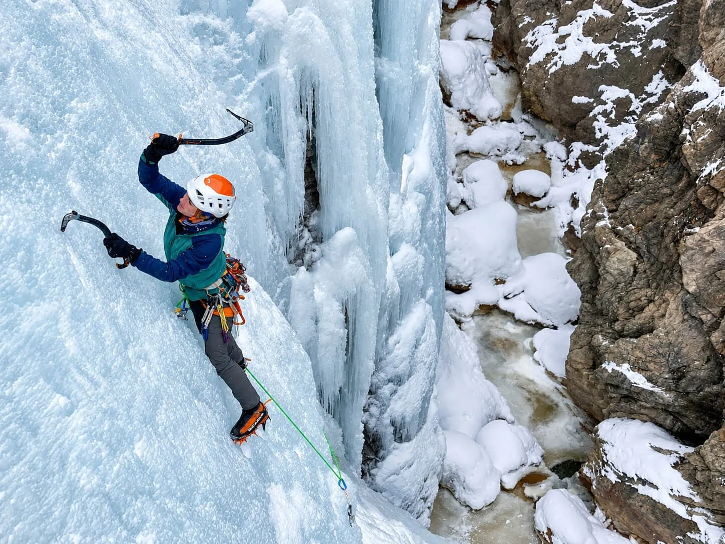 A guide leads a route in the Ouray Ice Park during an ice climbing course. She is wearing a white and orange helmet an teal jacket. She is swinging her ice tool.