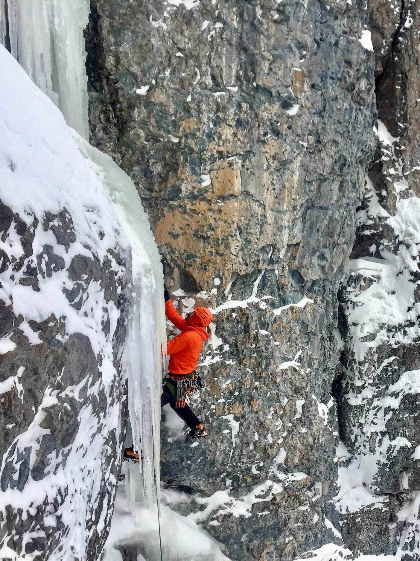 A person in a red jacket is lead climbing a mixed pitch of rock and ice with crampons and ice tools. He is swiging his ice tool into chandelier icicles coming down the corner above him.