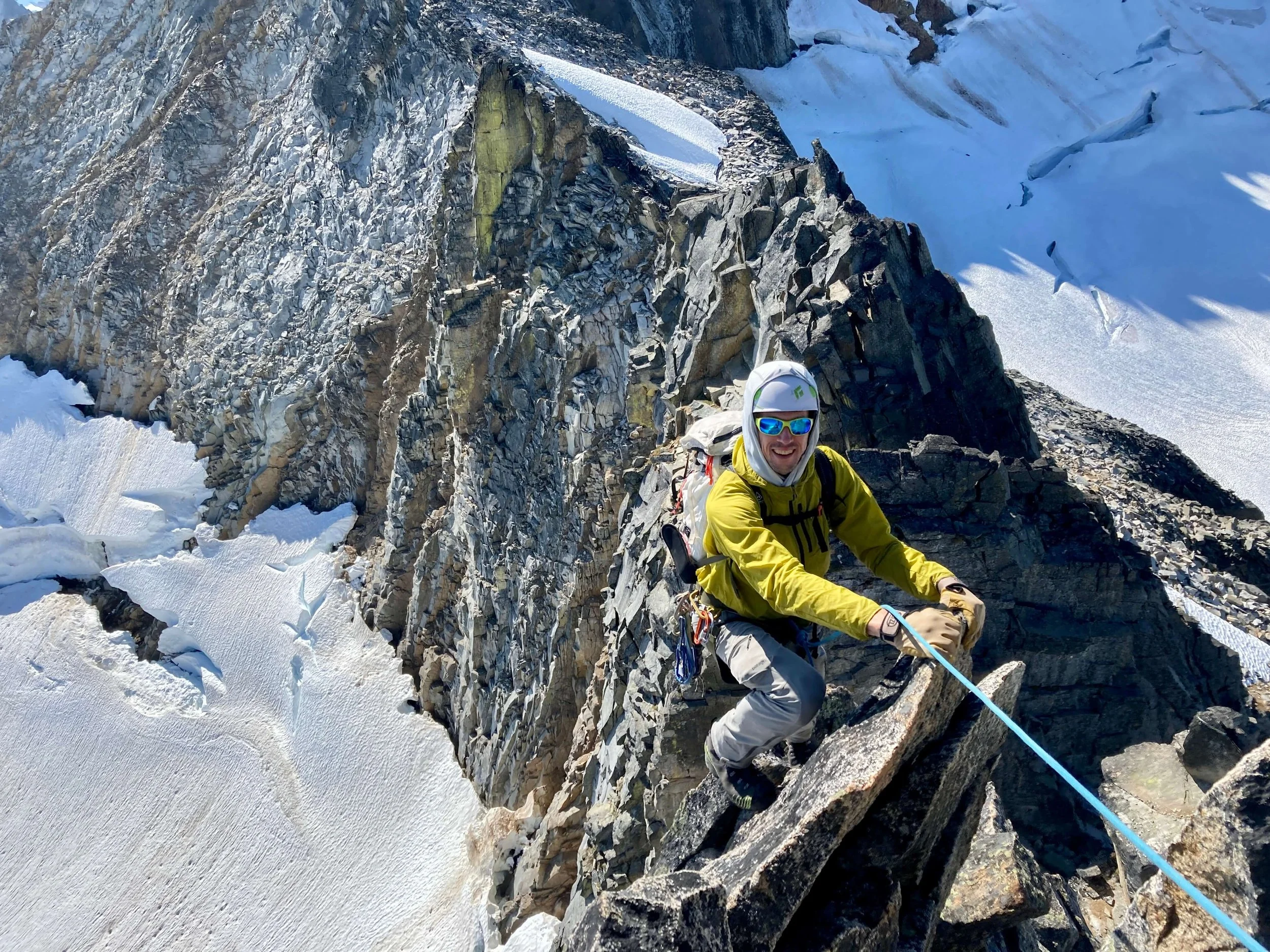 A climber in a yellow jacket is alpine climbing an exposed rocky ridge on Sharkfin Tower in Boston Basin with the Quien Sabe and Boston Glaciers below in the background.