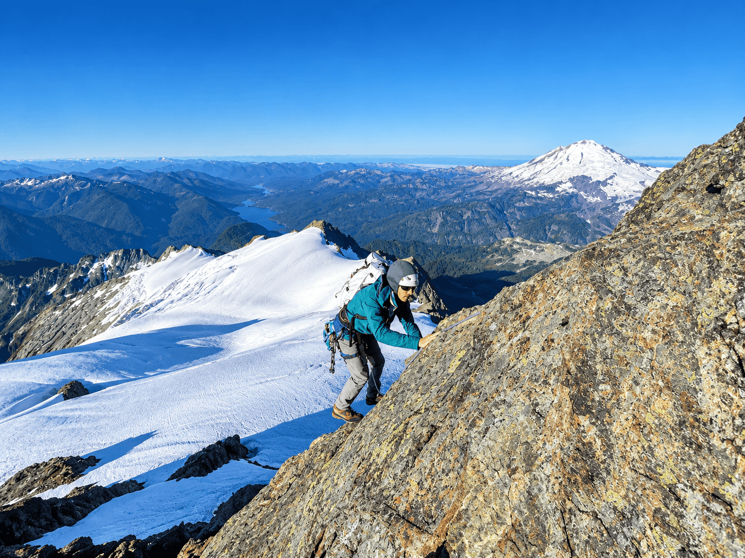 A climber in a green jacket ascends the South Ridge on Mount Shuksan's summit pyramid. Mount Baker rises in the background.