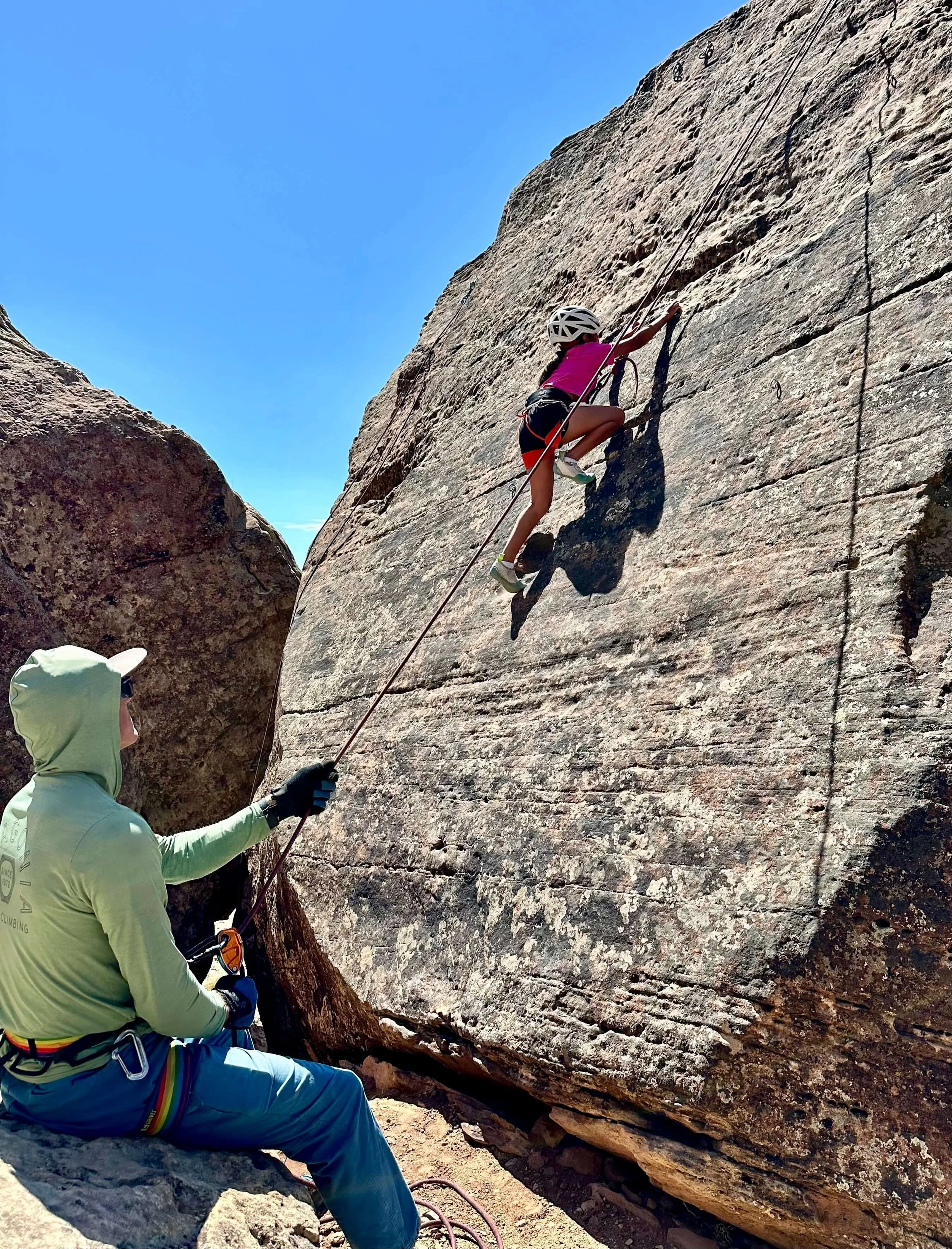 A young child in a pink shirt and white helmet is rock climbing at a crag in Grand Junction, being belayed by a guide in a green shirt.