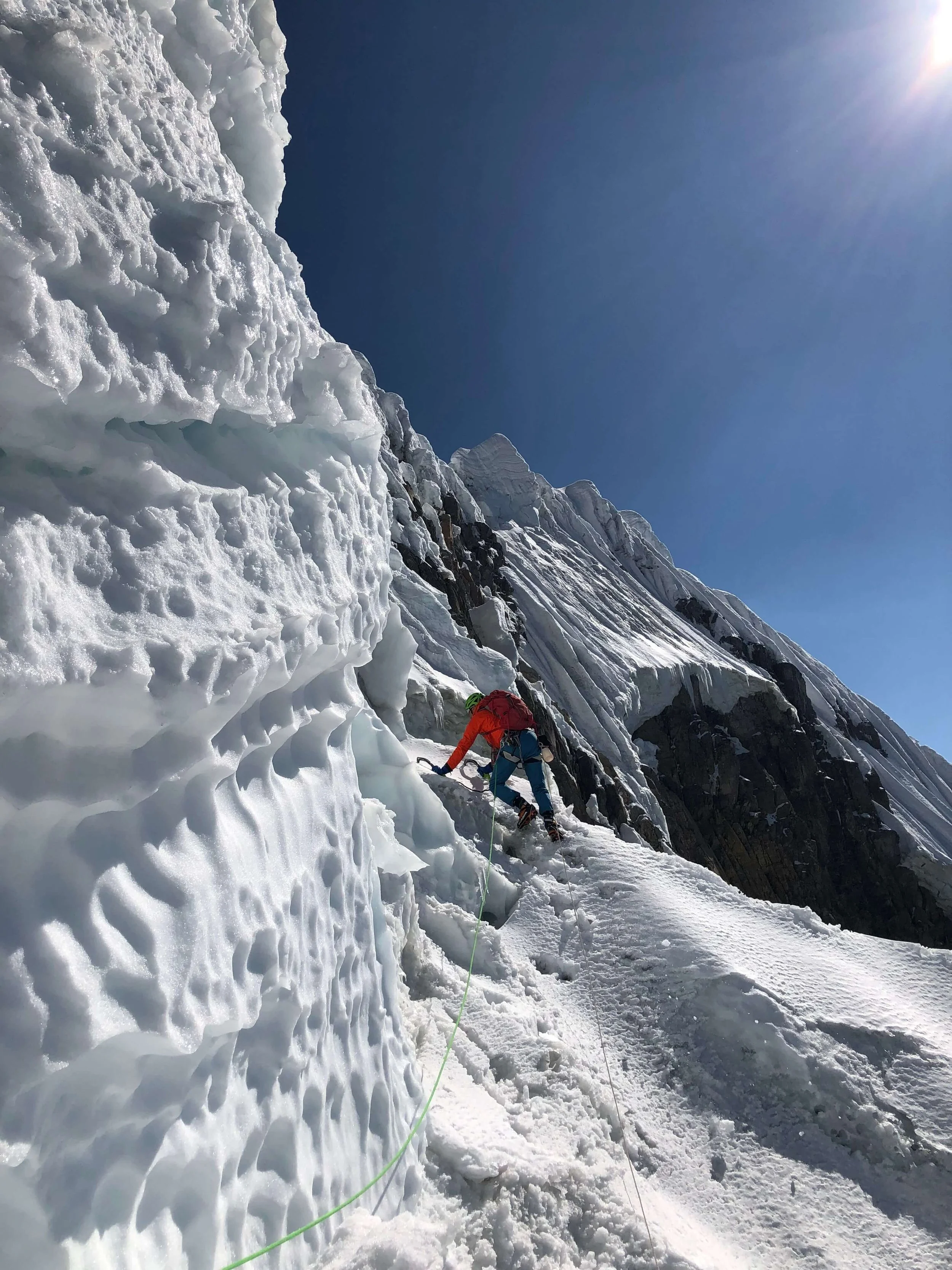 A climber ascending a steep, icy mountain face during a bright, sunny day.