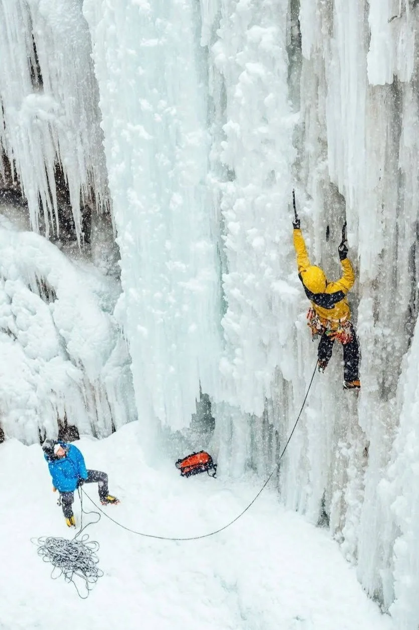 A climber in a yellow jacket is leading an ice climb with an overhanging start. He is being belayed from a woman in a blue jacket below.