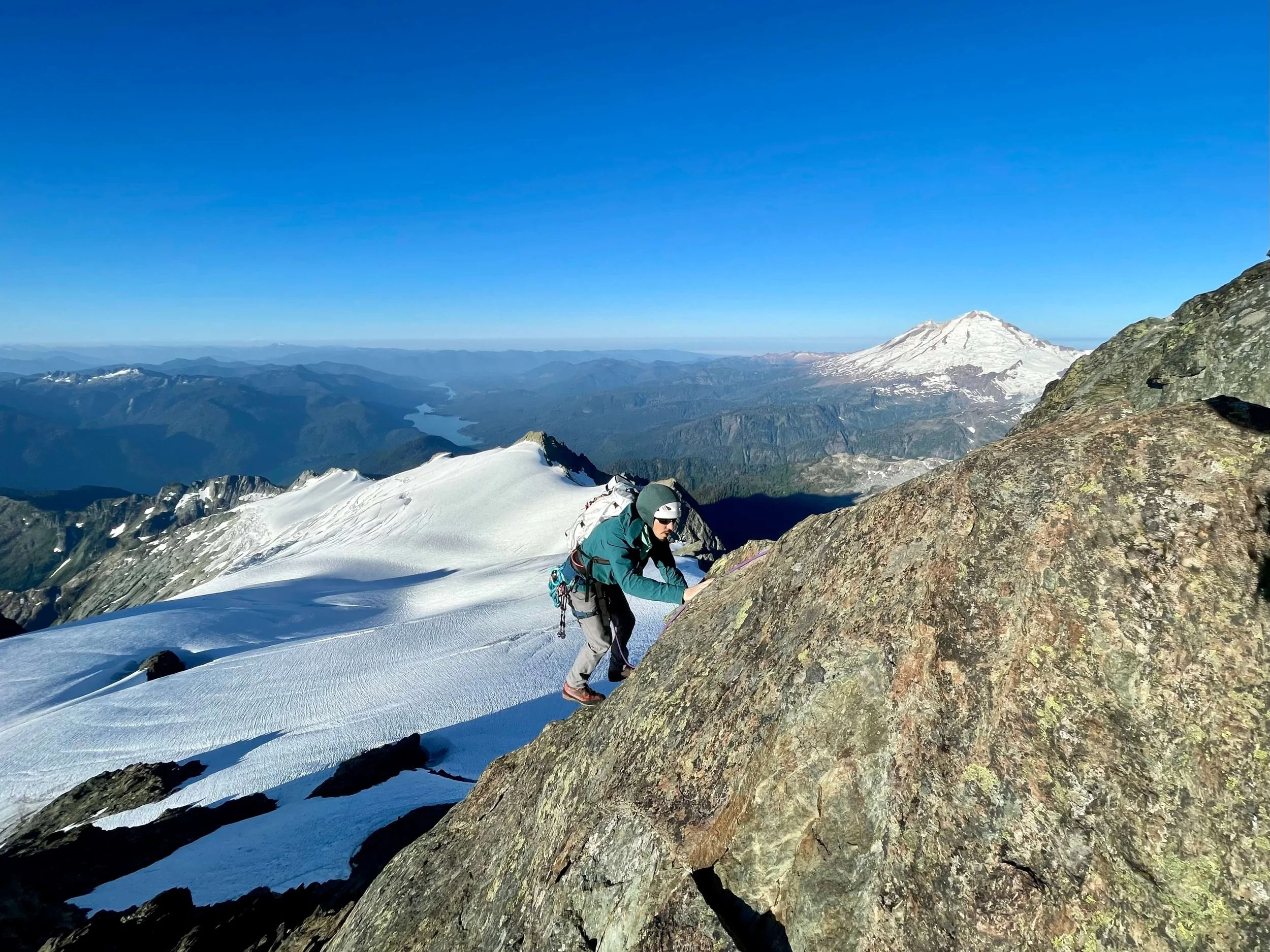 A climber in a green jacket ascends the South Ridge on Mount Shuksan's summit pyramid. Mnt Baker rises in the background.
