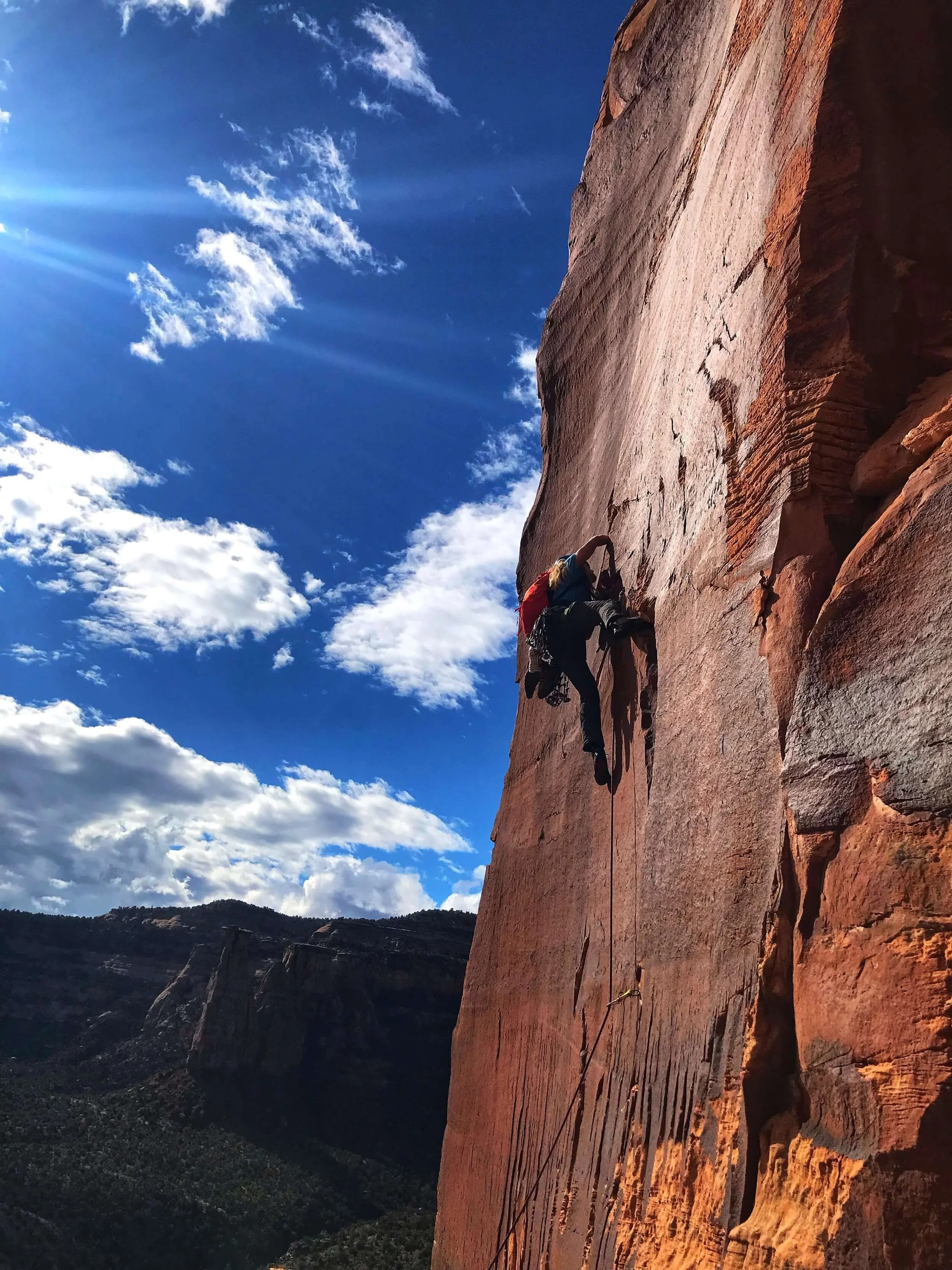 A silhouetted person is lead climbing a thin crack on a red sandstone desert tower in Colorado National Monument, set against a blue sky with clouds and distant mountains.