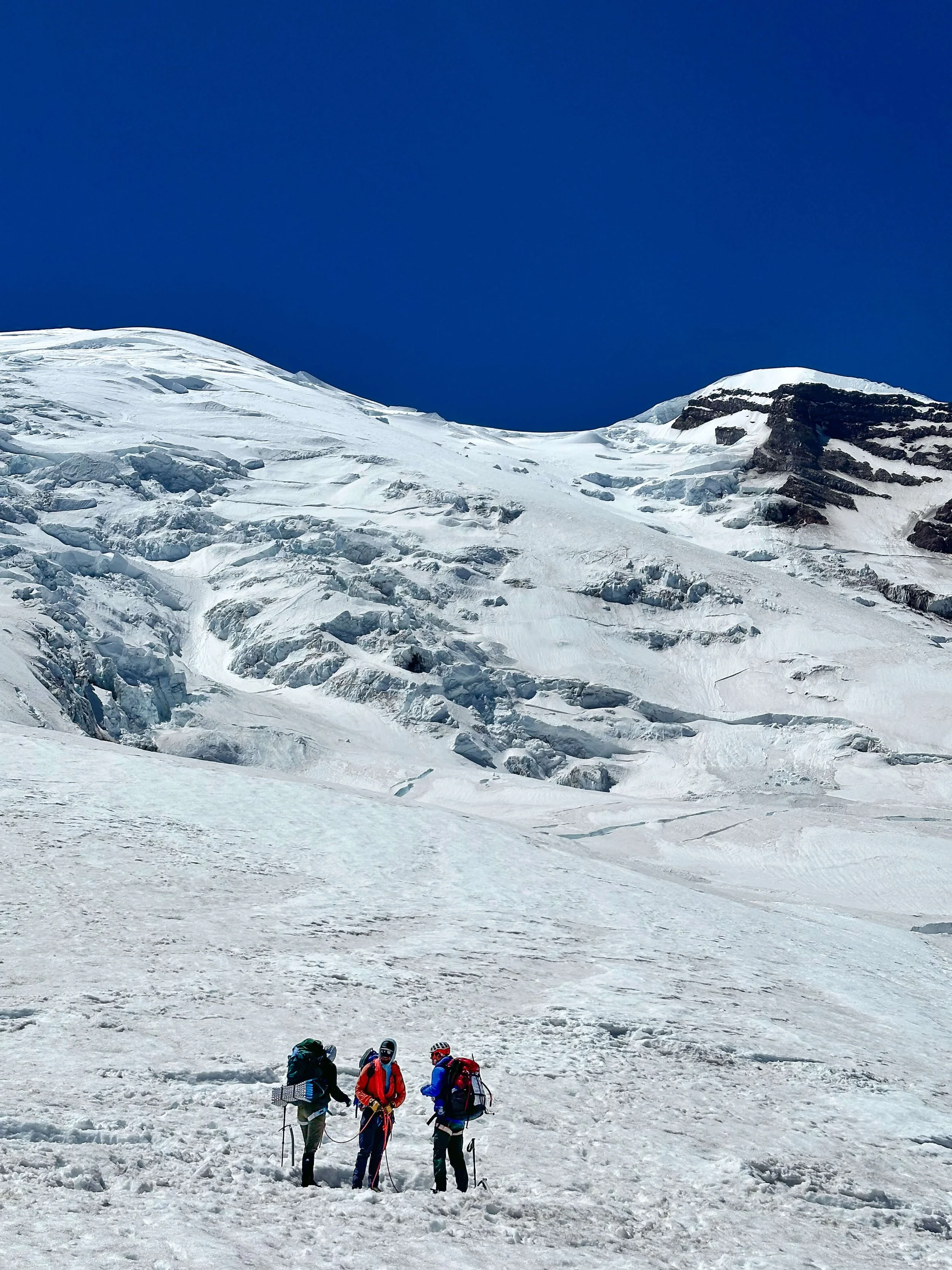 A group of three mountaineers stand below the large Emmons Glacier on Mount Rainier.