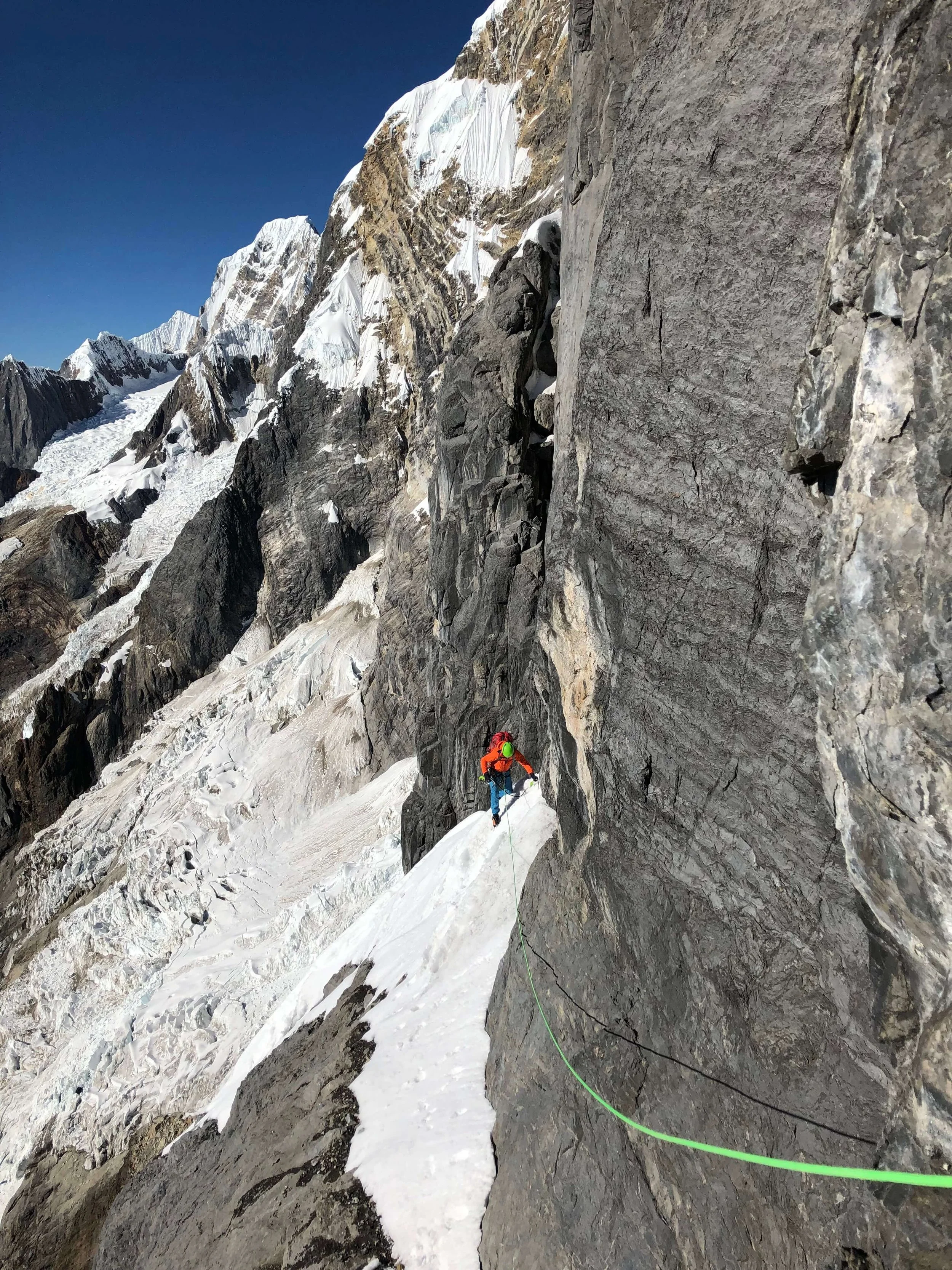 A climber in an orange jacket and green helmet traverses a steep snow face in the Peruvian Andes.