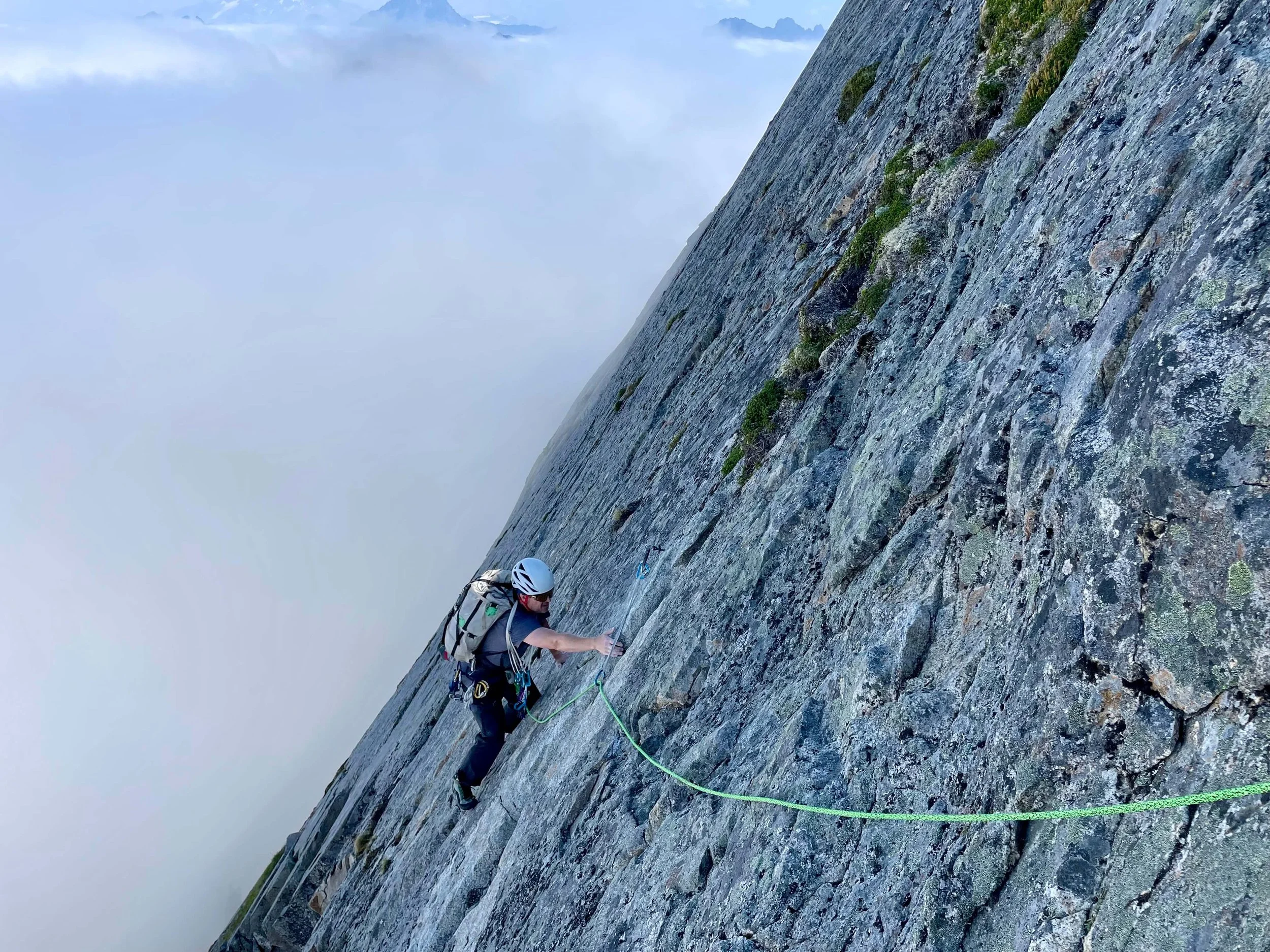 A rock climber in a white helmet is traversing a steep slabby face on the route Ragged Edges on Vesper Peak. A sea of clouds rises in the background.