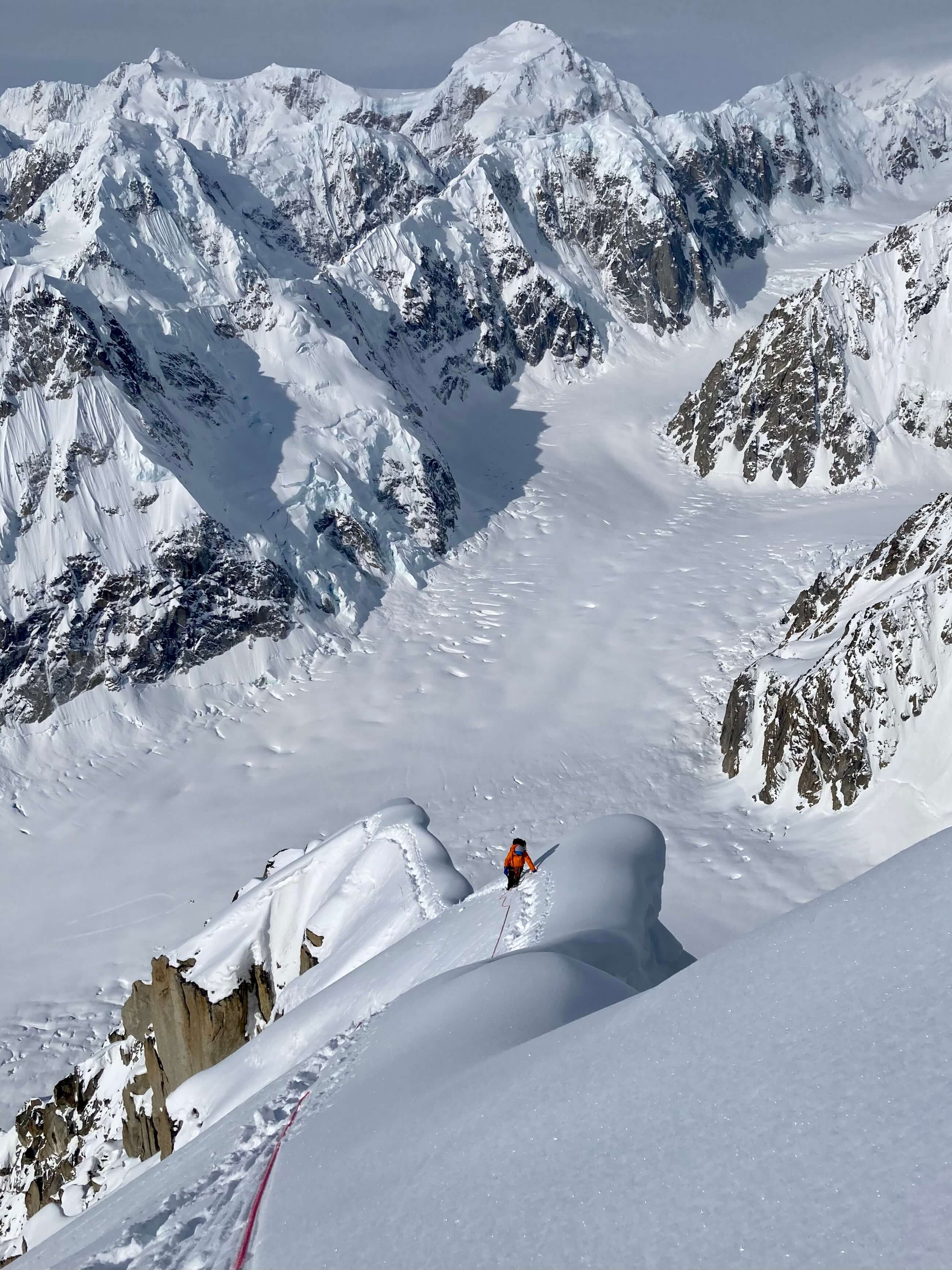 A climber in a red jacket is traversing a corniced ridge on Peak 11,300 in Alaska. The West Fork of the Ruth Glacier is below in the background.