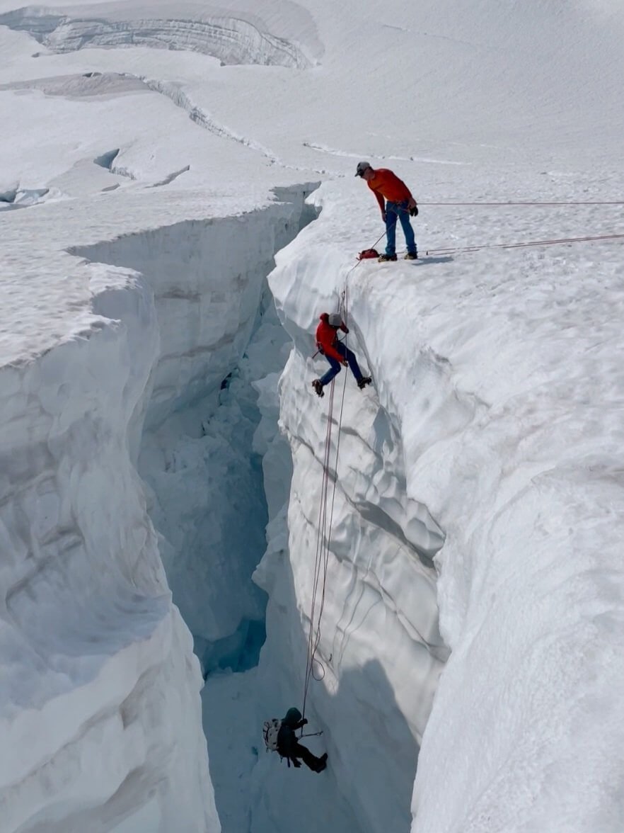 A climber is rappelling into a crevasse on a snowy glacier to practice rescue techniques while the guide looks on from above.