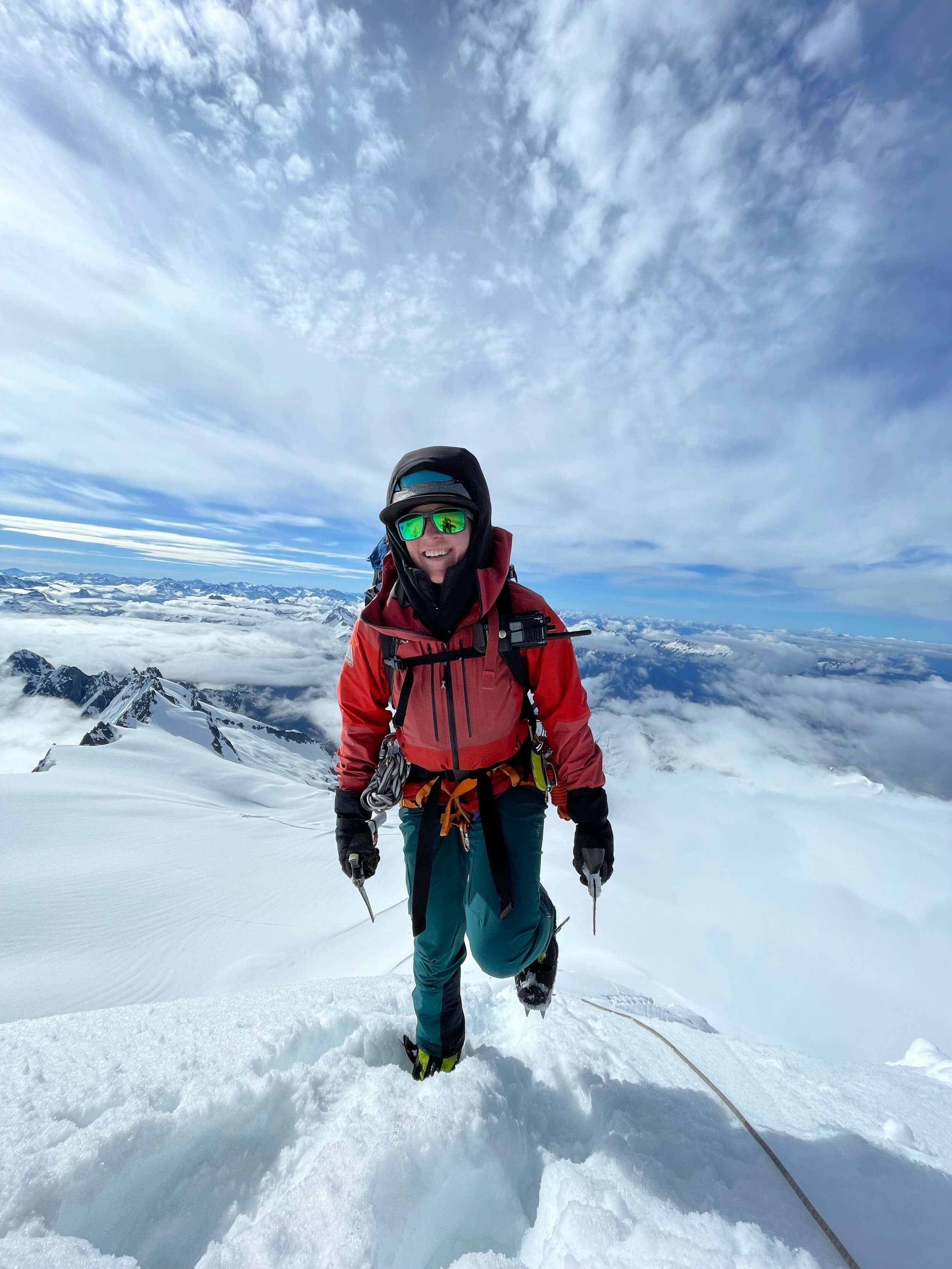 Climber wearing red jacket and teal pants climbs to the summit of snowy mountain. She is smiling with clouds and the Cascade mountains in the background.
