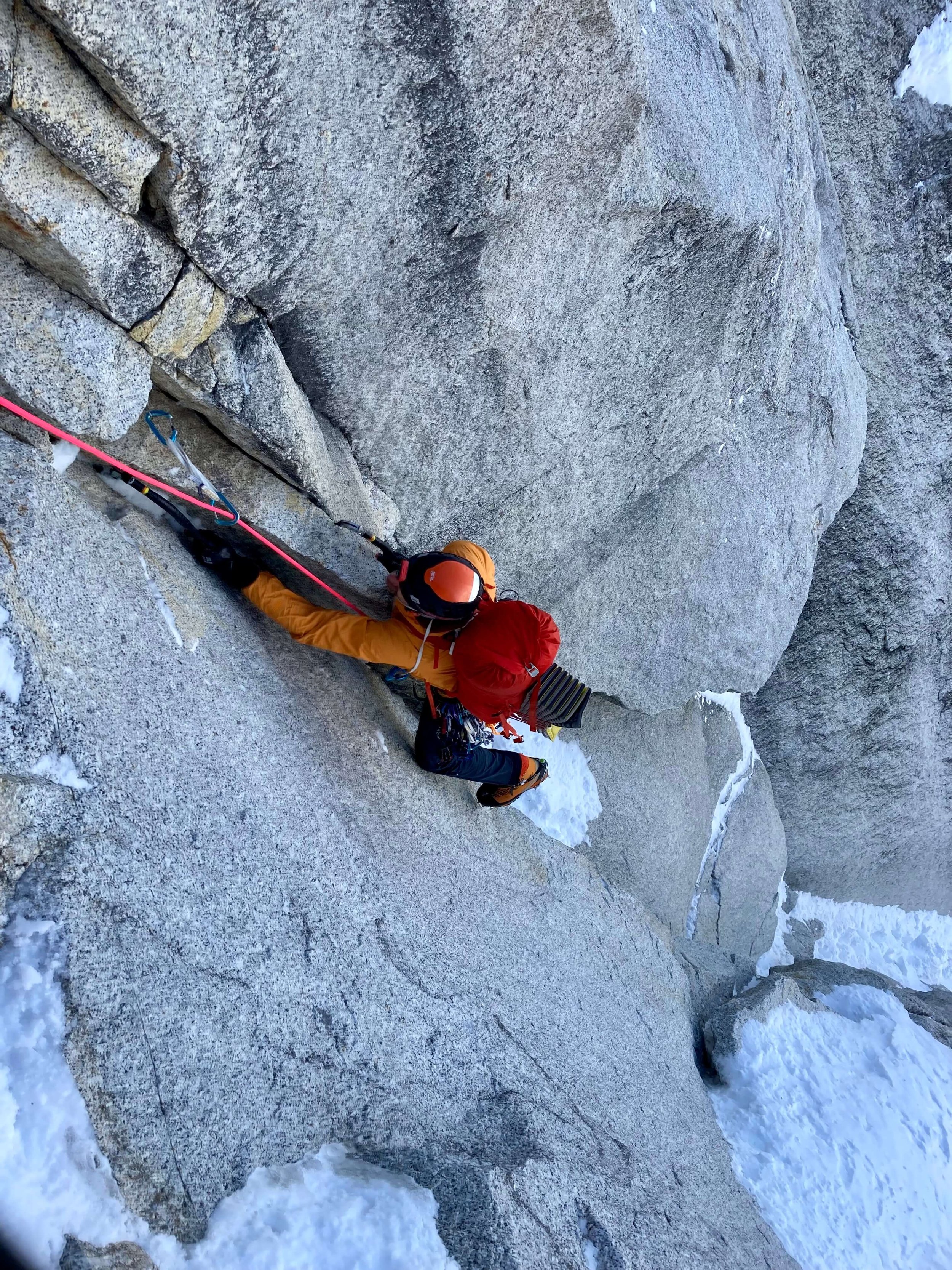 A climber in an orange jacket and red backpack is mixed climbing up a steep rock face on Mount Huntington's Harvard Route.