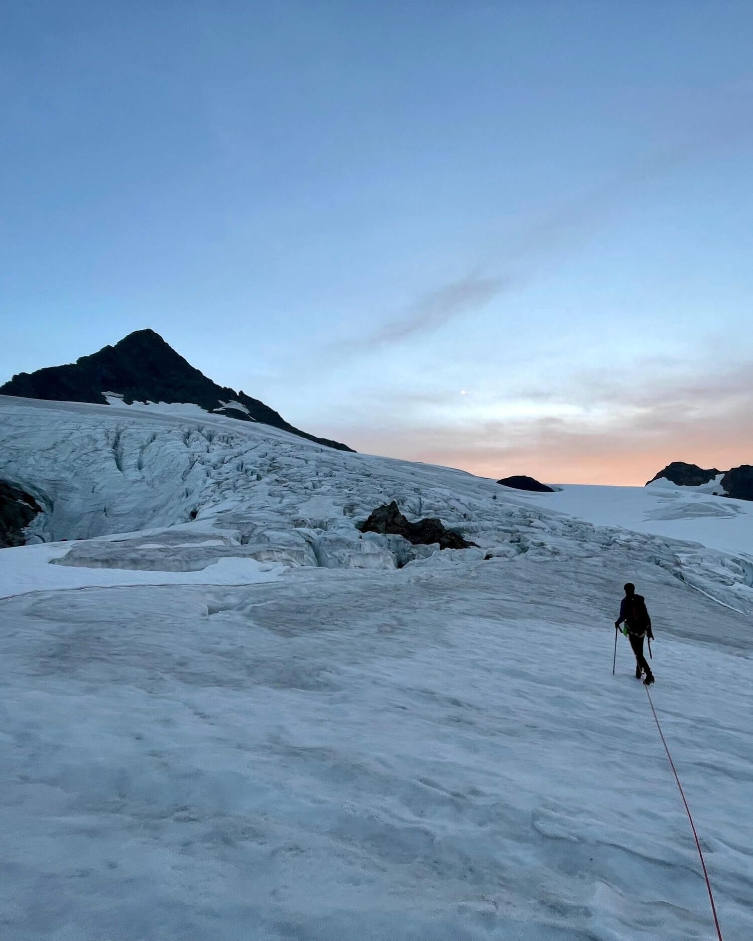 A person climbing a snowy mountain slope towards Mt. Shuksan's summit pyramid, with sunrise peeking out from behind the mountains.