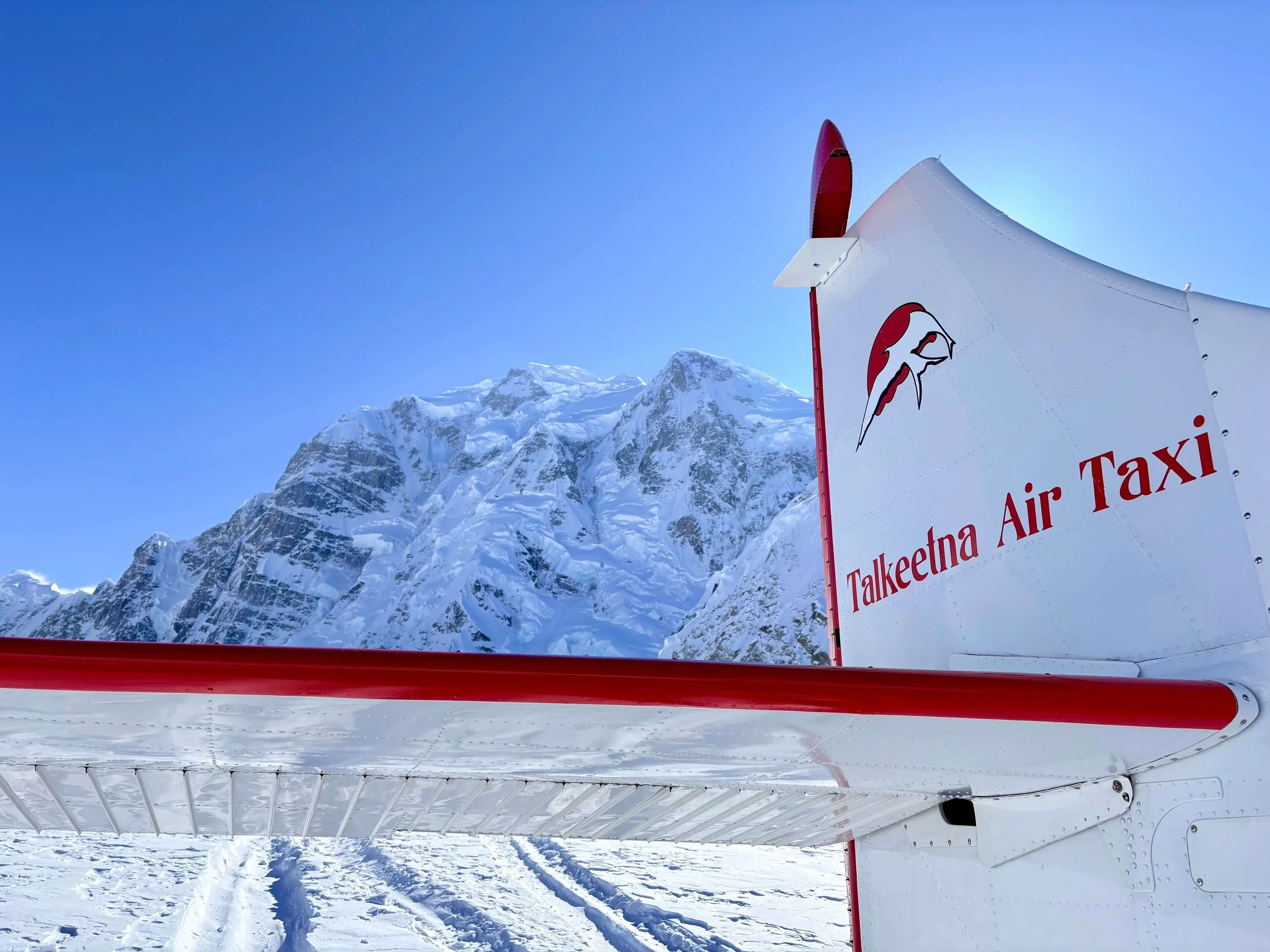 The tail of an airplane with the words "Talkeetna Air Taxi" sits on the Kahiltna Glacier with a snowy peak in the background.