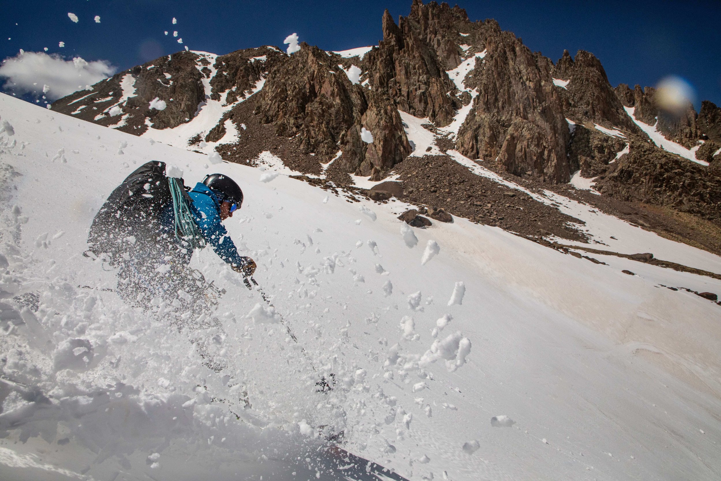 A skier in a blue jacket is skiing the steep snow slope below the Snake Couloir on Mount Sneffels. He is turning sharply and spraying powder into the air.