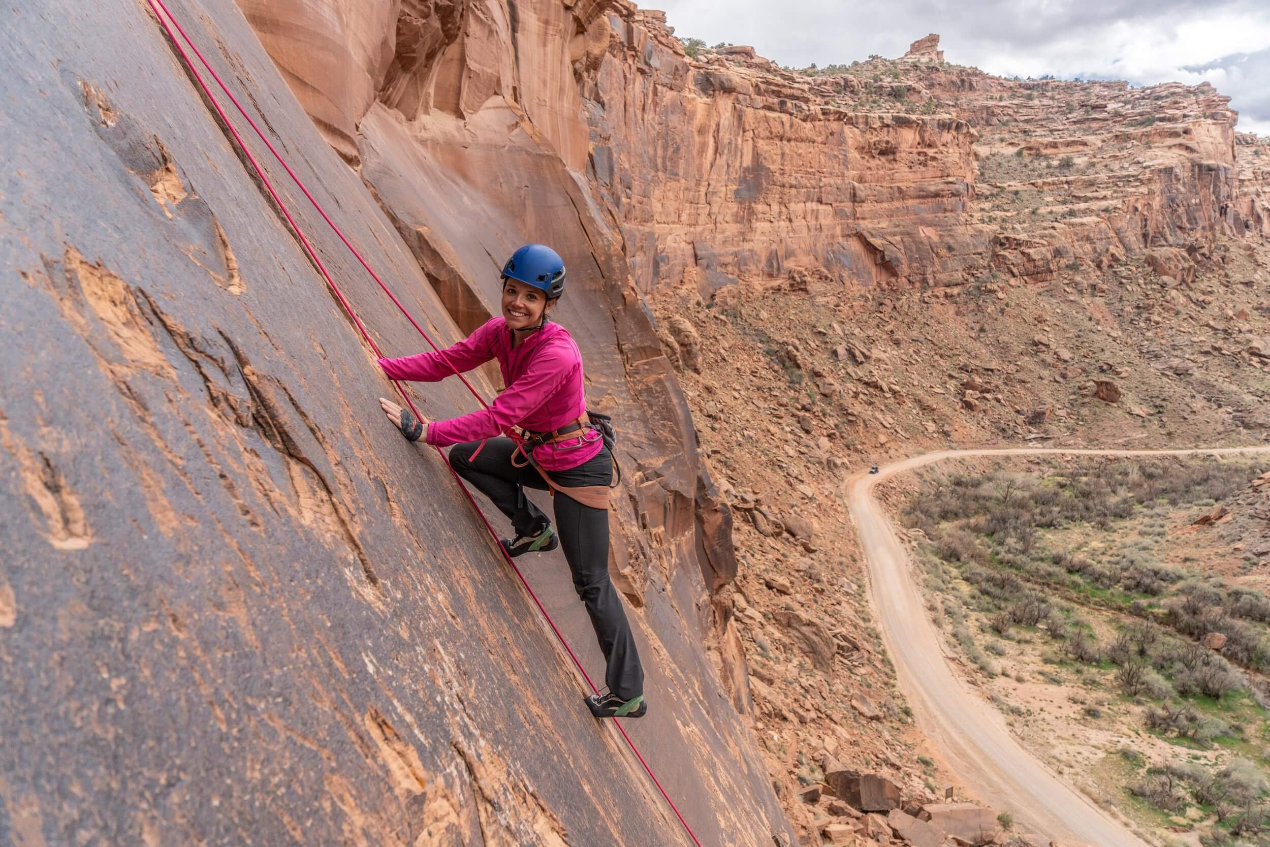 Woman rock climbing on a steep cliff in Moab's desert landscape with a winding dirt road below.