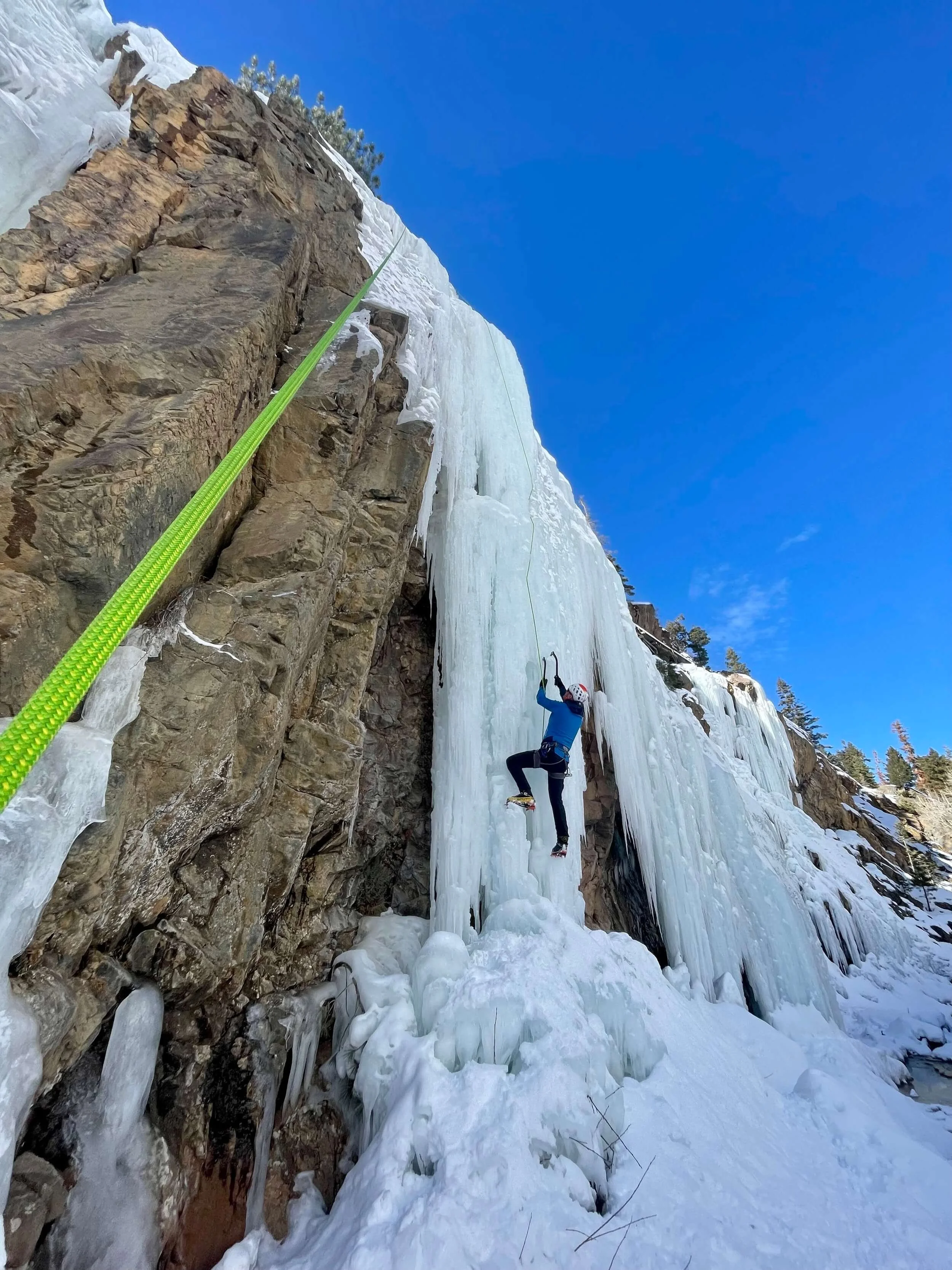Person ice climbing on a frozen waterfall with ice tools and crampons, secured by a bright green climbing rope, on a clear winter day.