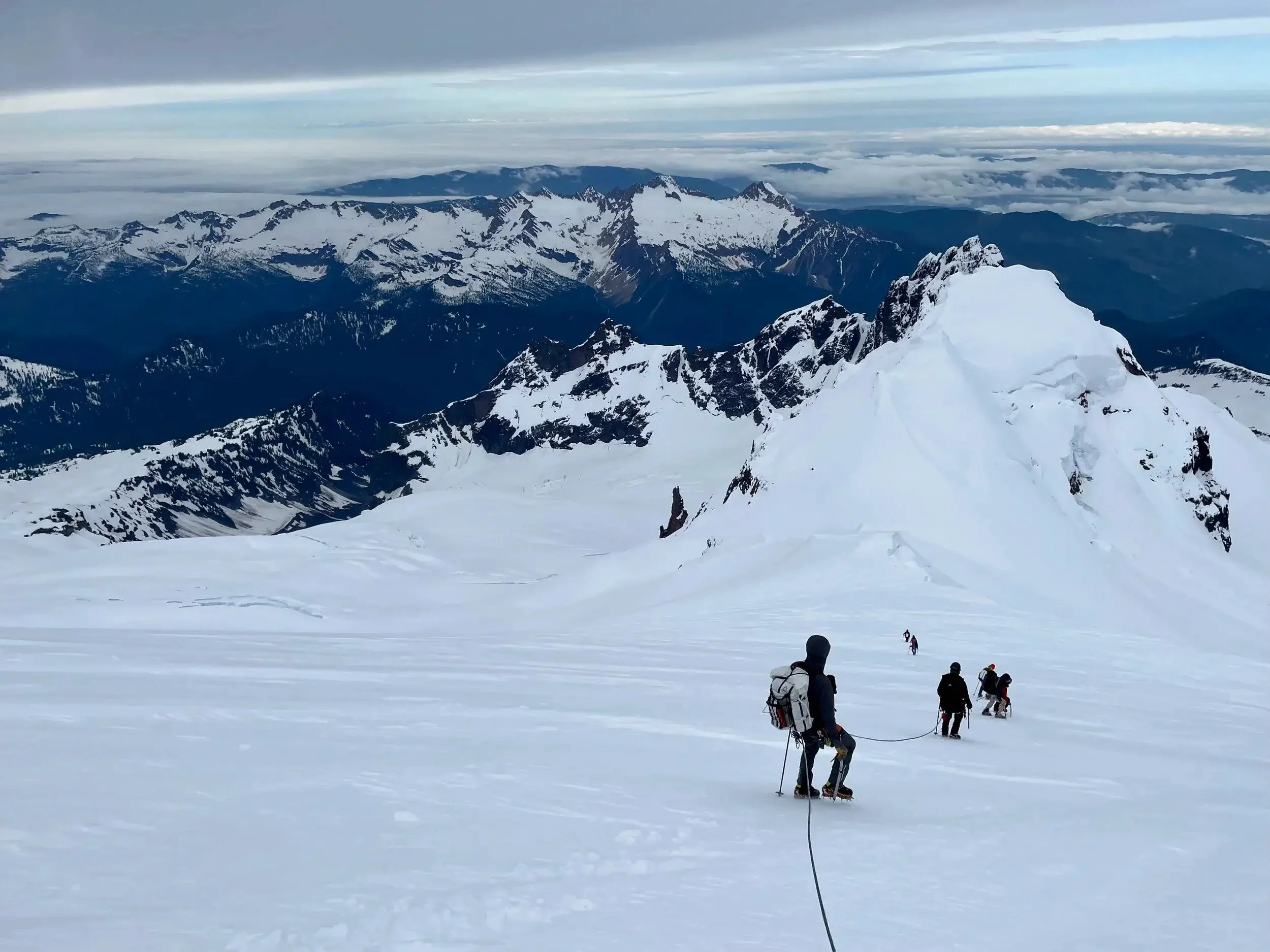 A group of mountaineers descending from the summit of Mount Baker during an early spring ascent. They are down climbing a steep, snowy ridge with glaciers on both sides.