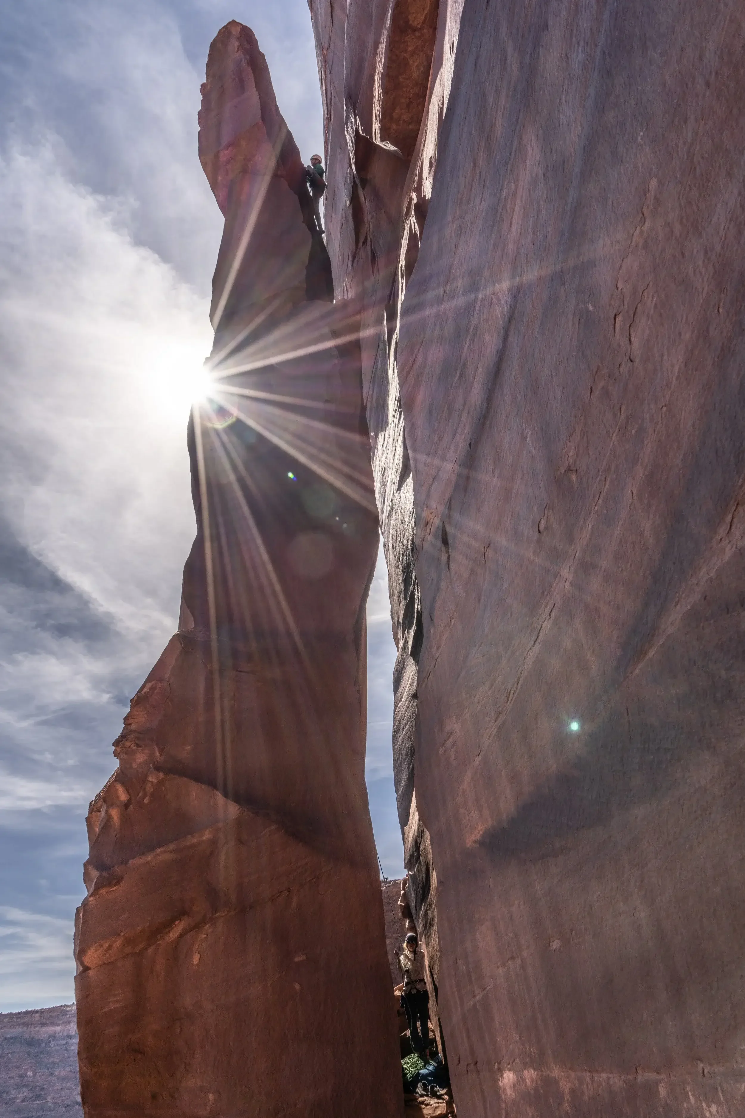 A rock climbing guide is leading up a desert tower. She is climbing up a chimney with the sun peeking out from the side of the tower.