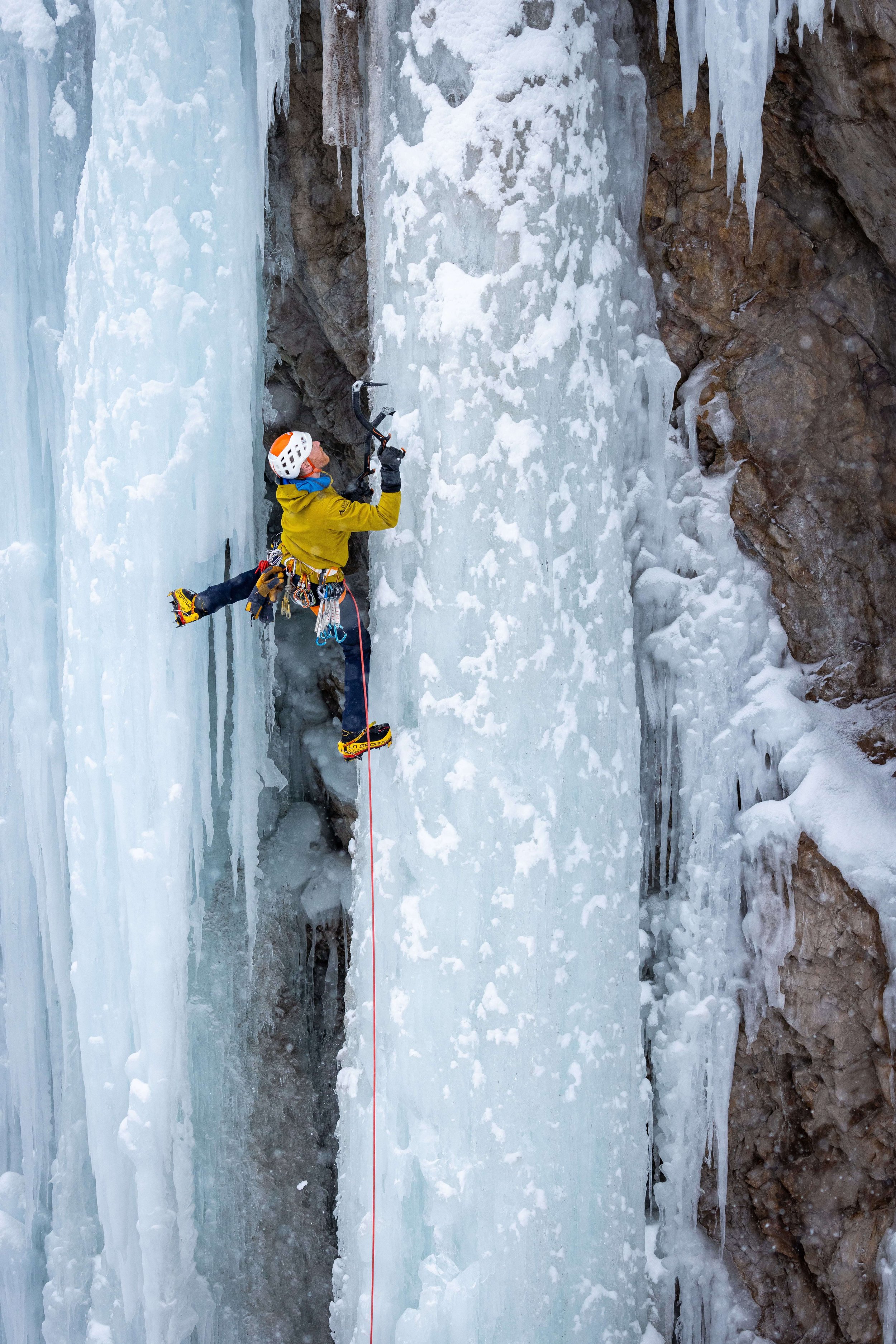 An ice climber in a yellow jacket is stemming across two steep pillars of ice and swinging his ice tool into the right side pillar while leading a route in the Ouray Ice Park.