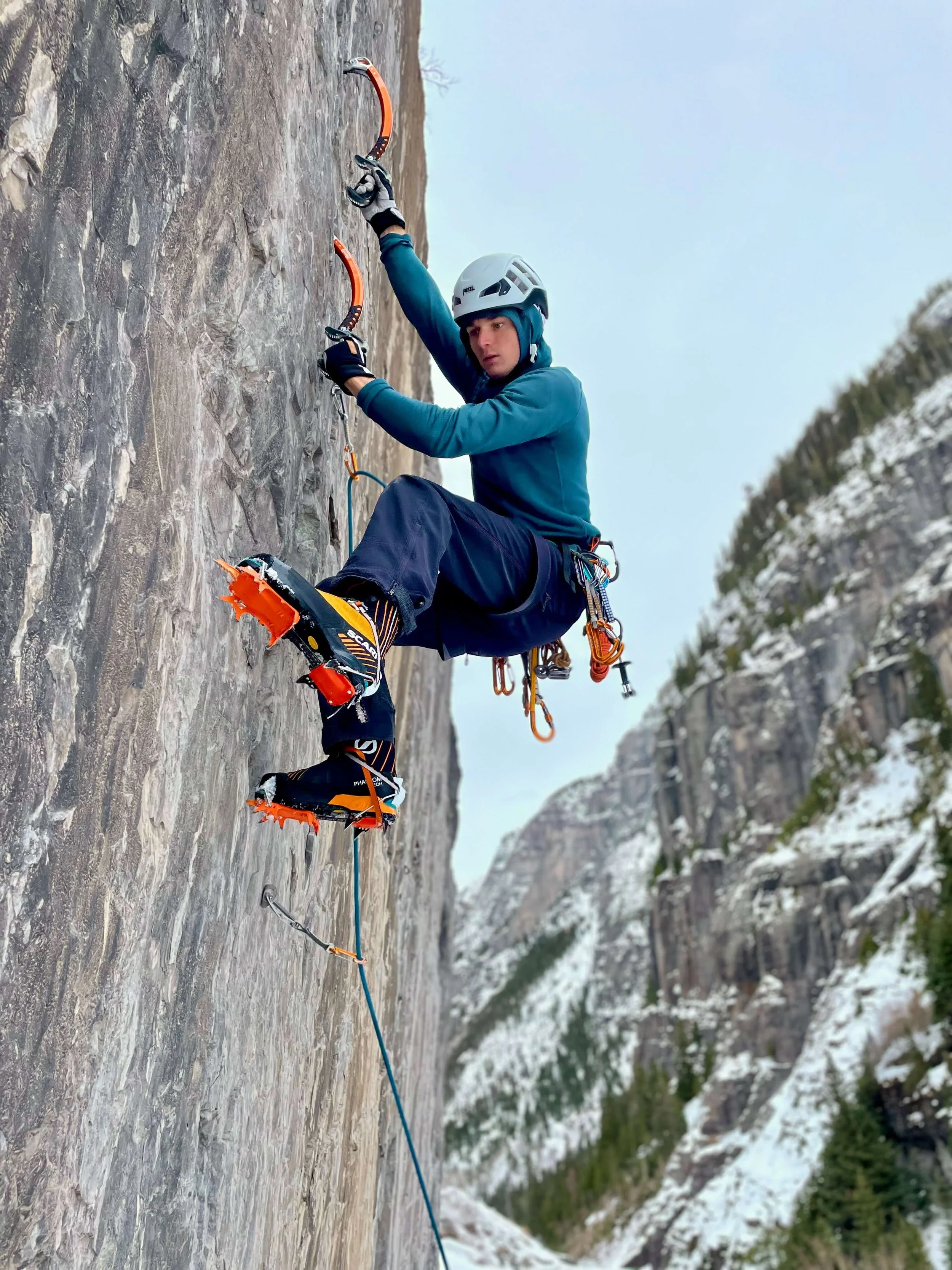 A climber in a blue jacket is leading a mixed route on Camp Bird Road. He is placing his crampons on a small rock foothold.
