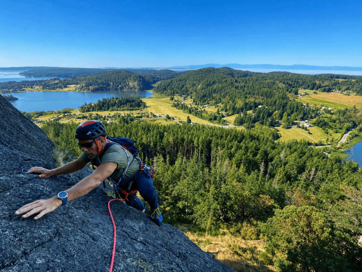 A climber in a green t-shirt is reaching for the next handhold on the route Zig Zag at Mount Erie. Several lakes and the Pacific Ocean are visible in the background.