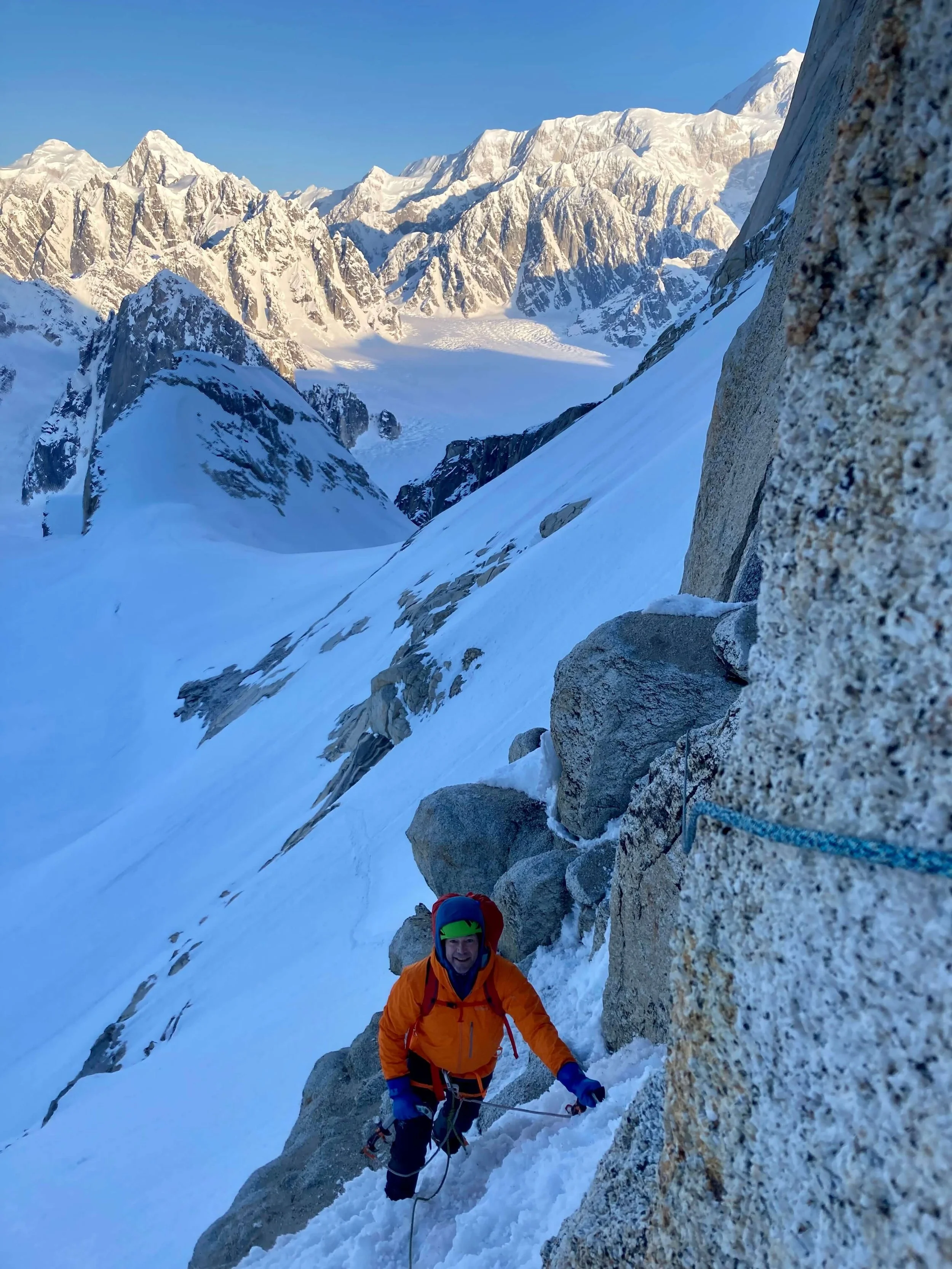 A climber in an orange jacket is traversing across the mixed rock and snow pitch at the start of Ham and Eggs on the Moose's Tooth.