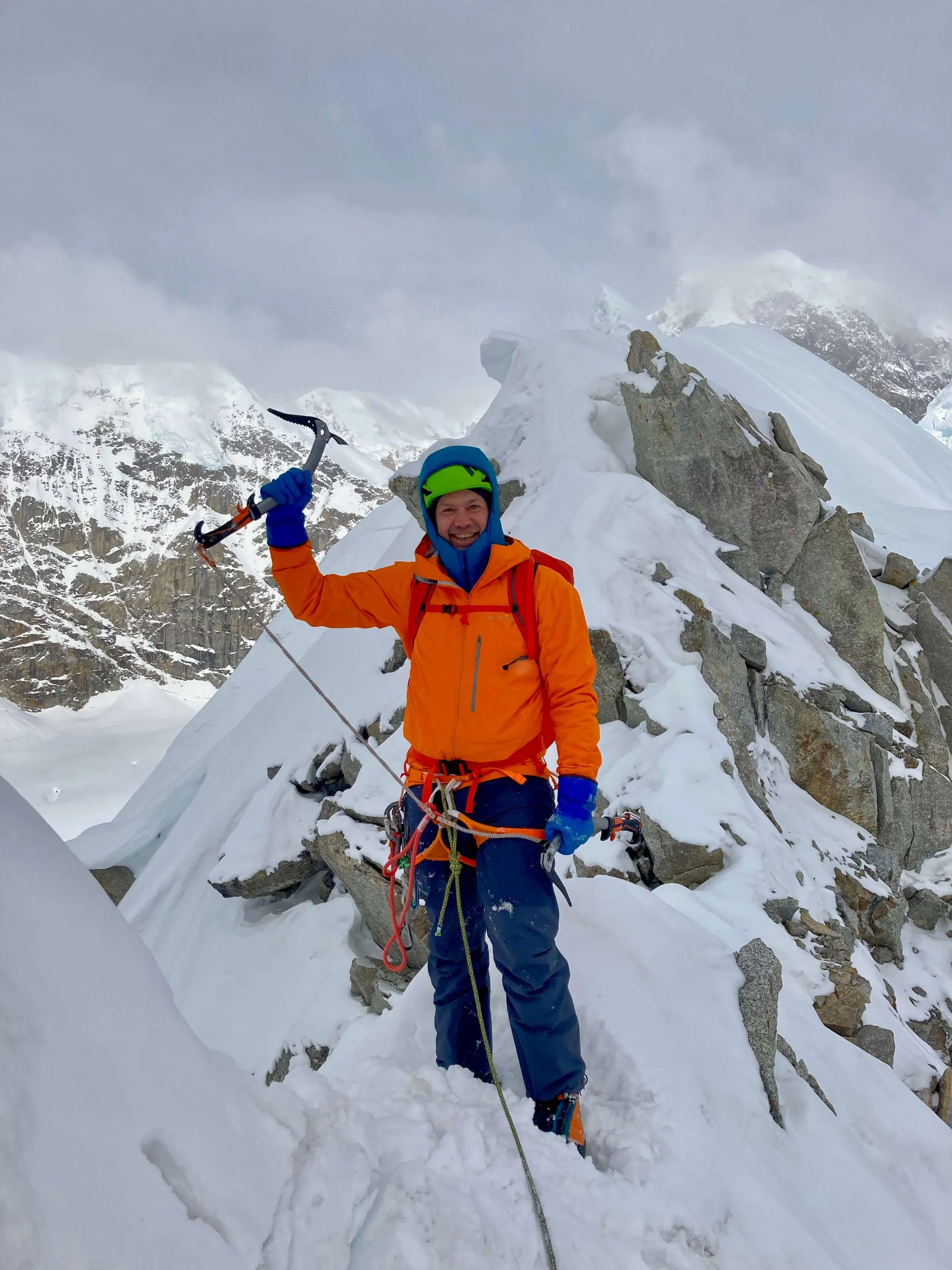 A climber in an orange jacket is holding an ice axe above his head on the summit of Mount Hunter in Alaska with a snowy ridge in the background.