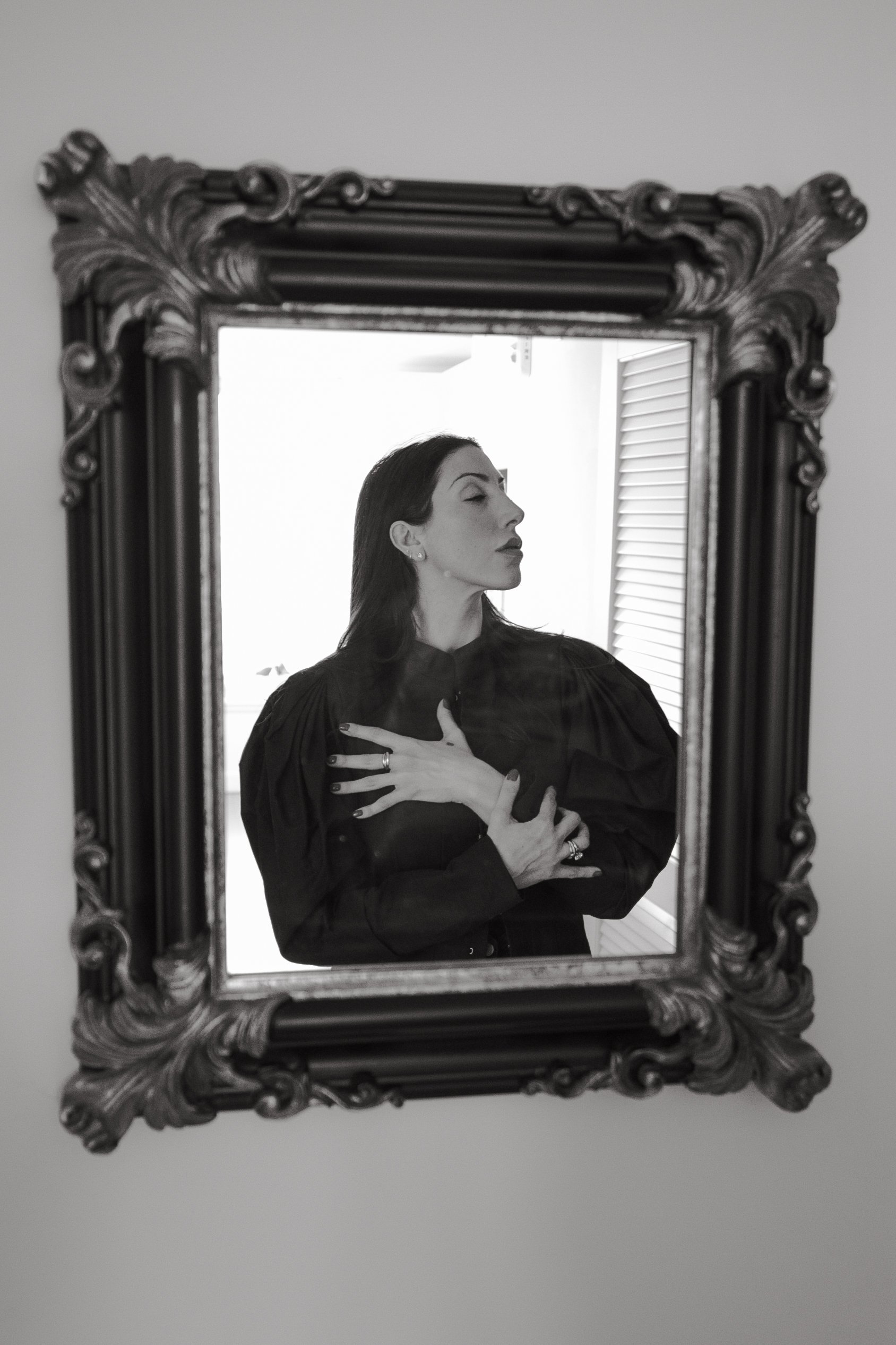 Black and white photograph of a woman in a dark dress with puffed sleeves, standing in front of a mirror and touching her chest with her hands crossed. The ornate frame of the mirror surrounds her reflection.