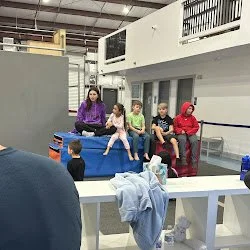 A group of six people, including children and adults, sitting on a blue bench in a parkour park, with some items on a table in the foreground.