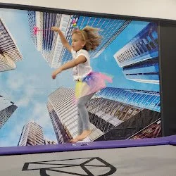 A girl jumping on an indoor trampoline with a cityscape background on the screen.