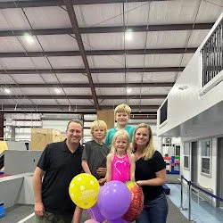 Group of five people, two children and three adults, standing inside a parkour gym, holding colorful balloons.