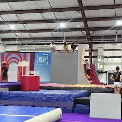 Children playing on a parkour trampoline with a foam pit and padded mats inside a gymnasium.