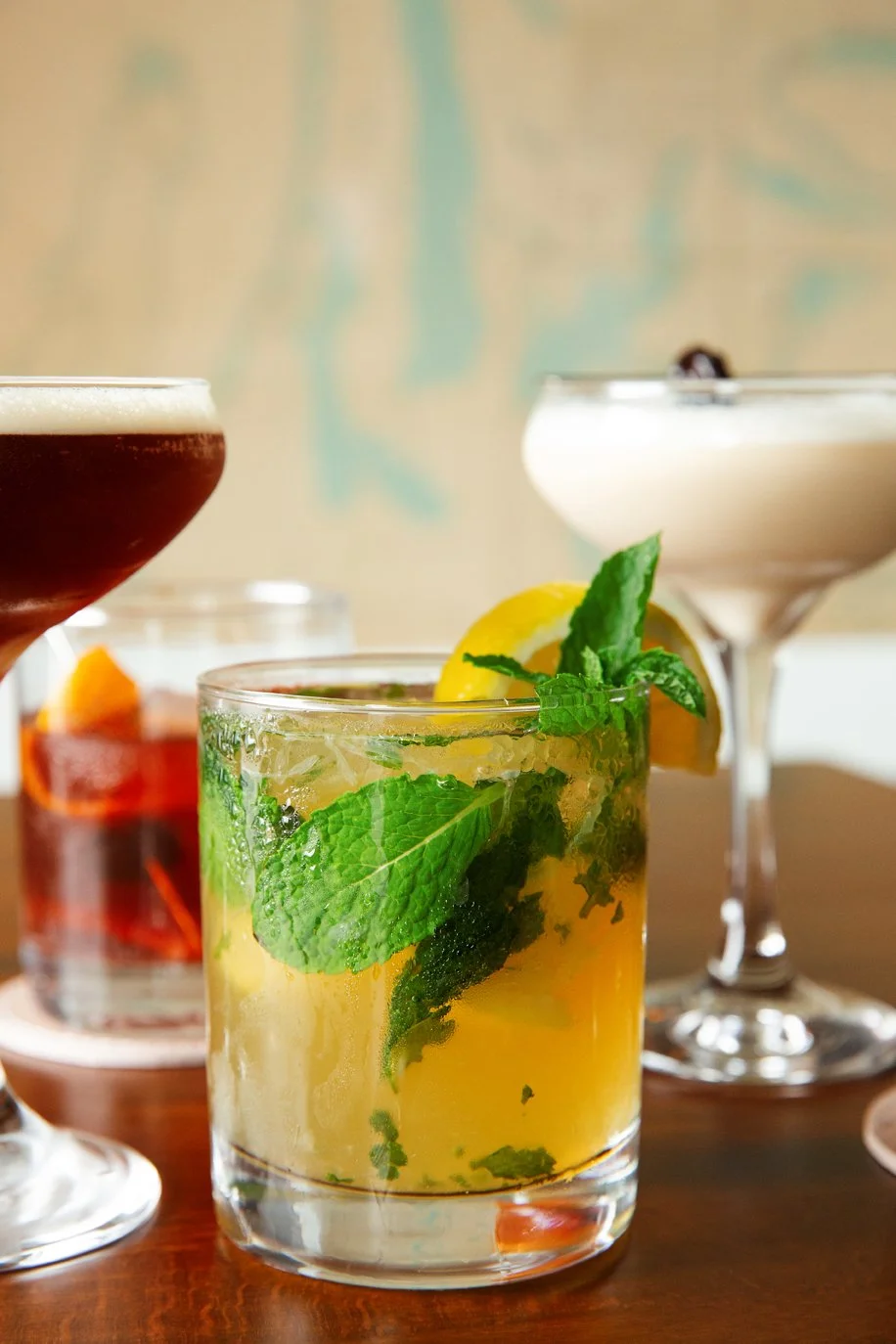 A glass of mojito cocktail with mint leaves, lemon wedge, and ice, alongside two other cocktails in the background on a wooden surface.