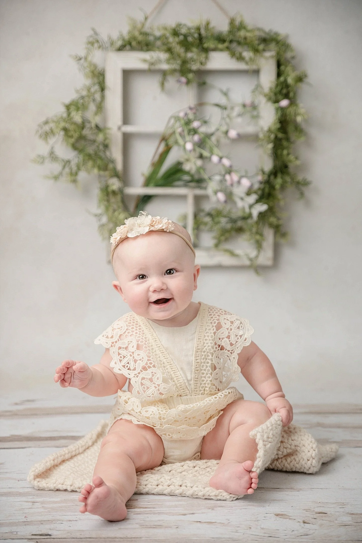 A baby girl sitting on a white wooden floor, wearing a cream lace outfit and a pink headband with floral accents, smiling at the camera with a backdrop of greenery and a white decorative frame.