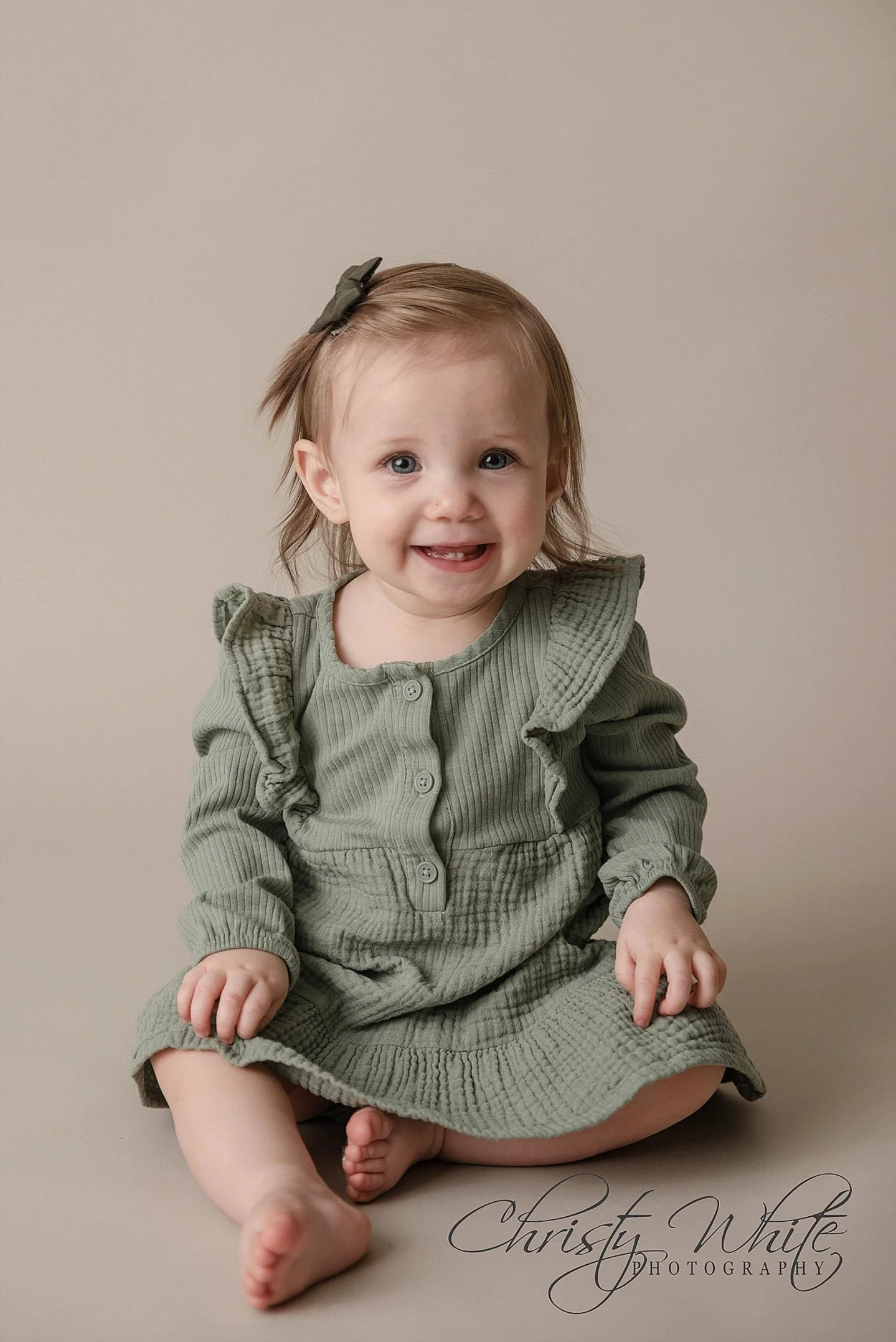 A young girl with light brown hair and a headband sitting on the floor, smiling at the camera, wearing a green dress with ruffles and buttons.