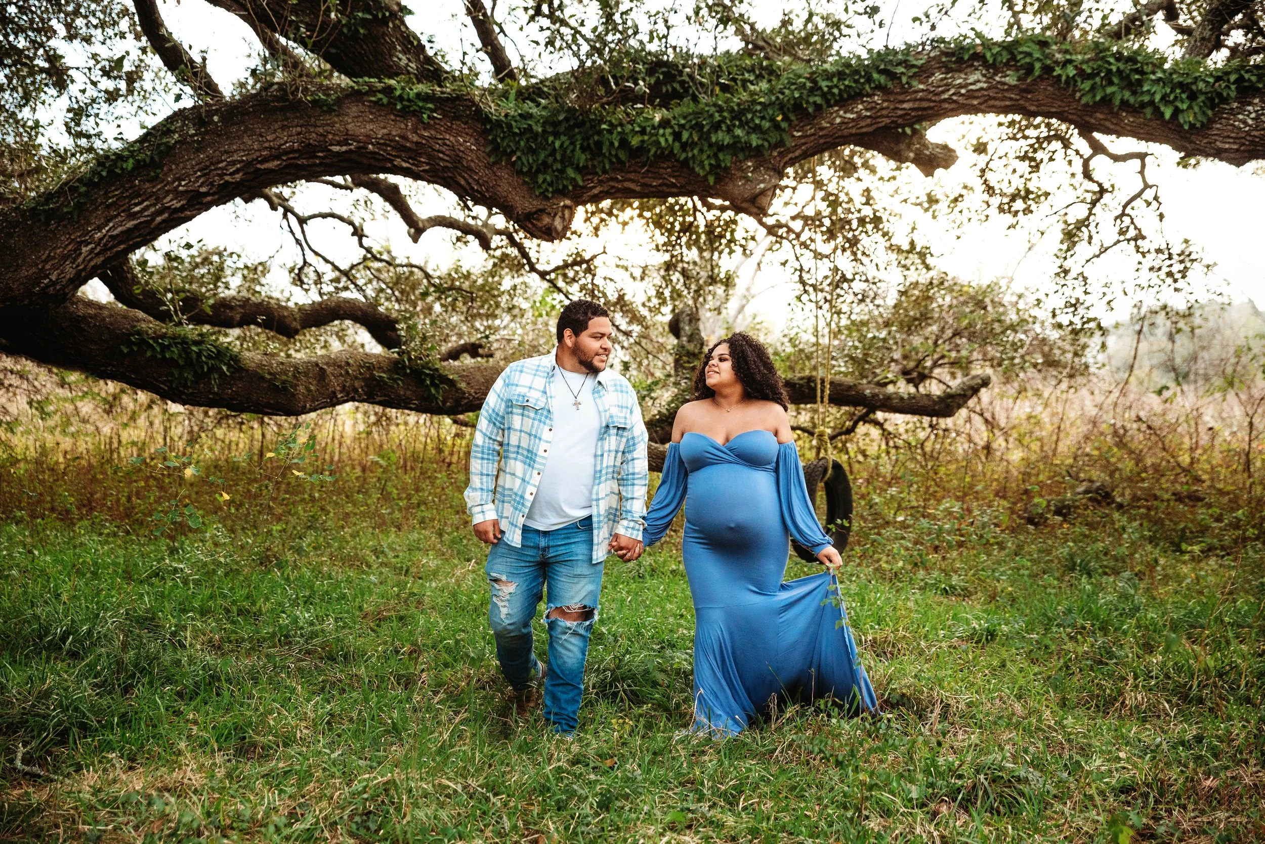 A pregnant woman in a blue dress holding hands with a man in a plaid shirt and ripped jeans, walking in a grassy outdoor area with a large tree and swing in the background.