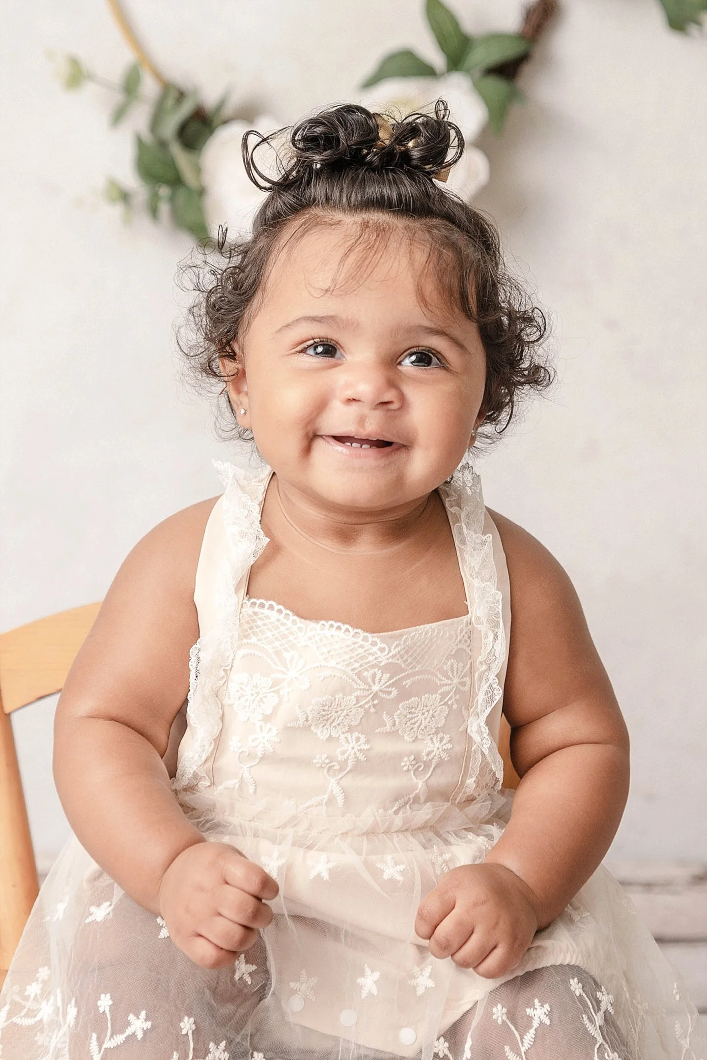 A smiling toddler girl with curly hair styled in a top bun, wearing a cream-colored lace dress, sitting on a wooden chair, with green leaves and white flowers in the background.