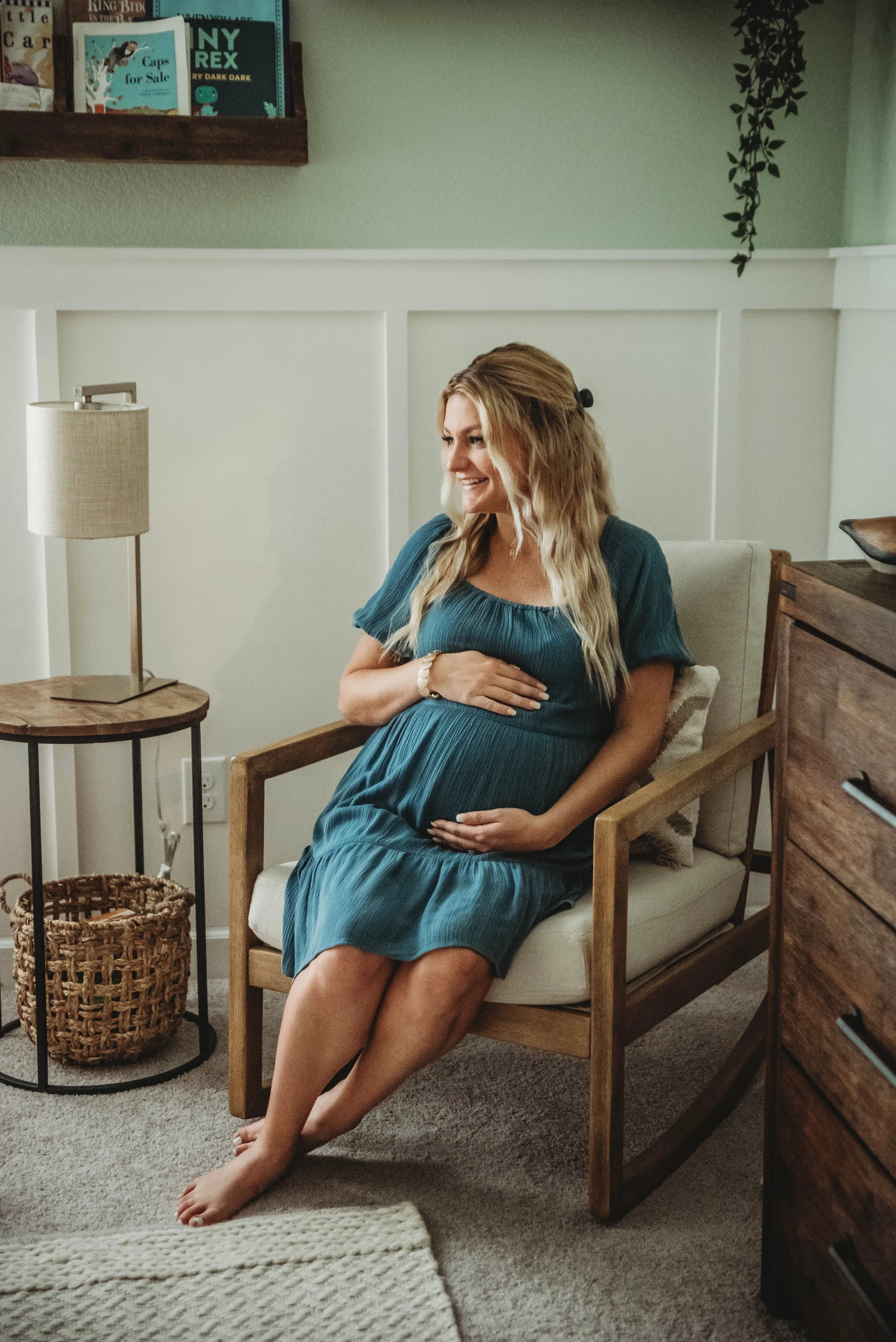 Pregnant woman sitting in a wooden chair indoors, smiling and holding her belly, next to a small table with a lamp, and a basket underneath, with books on a shelf above.