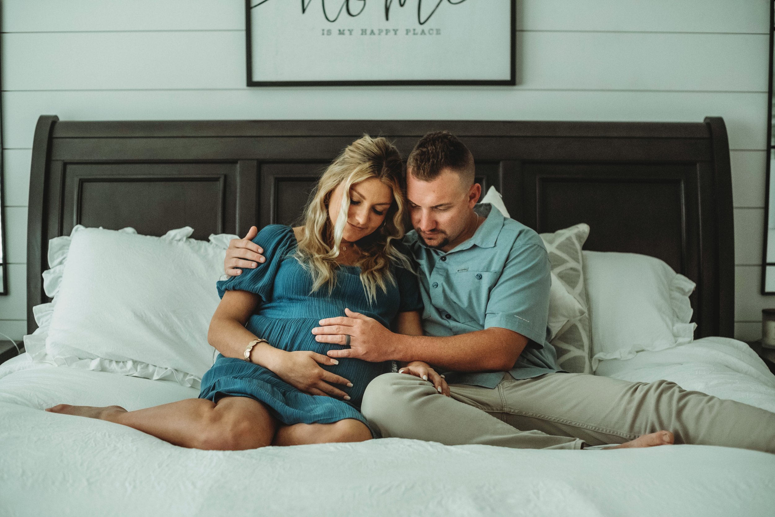 A pregnant woman and her partner sitting on a bed, touching her baby bump and looking down.
