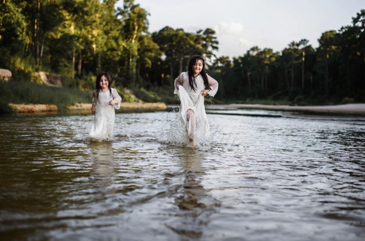 Two young girls wearing white dresses run through a shallow river, splashing water as they enjoy a sunny day outdoors, with trees and a partly cloudy sky in the background.