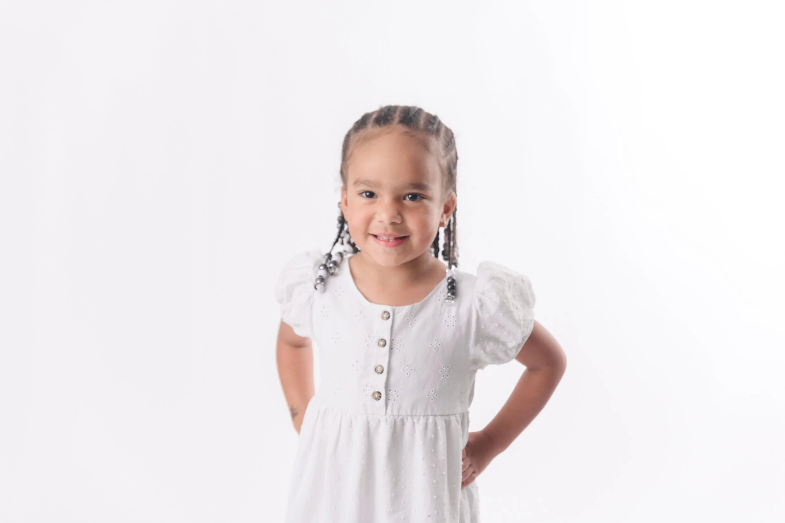 Young girl with braided hair wearing a white dress, smiling with hands on hips against a plain white background.