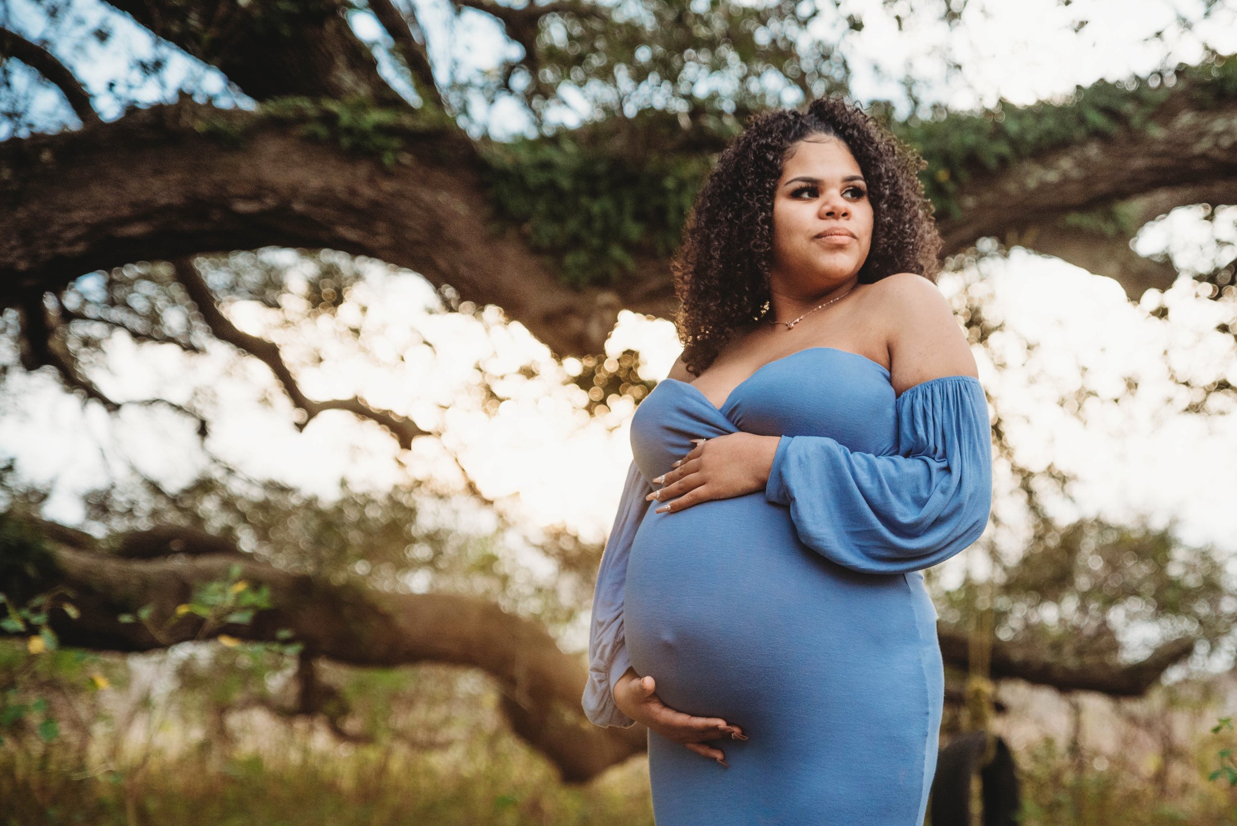 A pregnant woman in a blue off-shoulder dress standing outdoors near a large tree, with a patterned sky background.