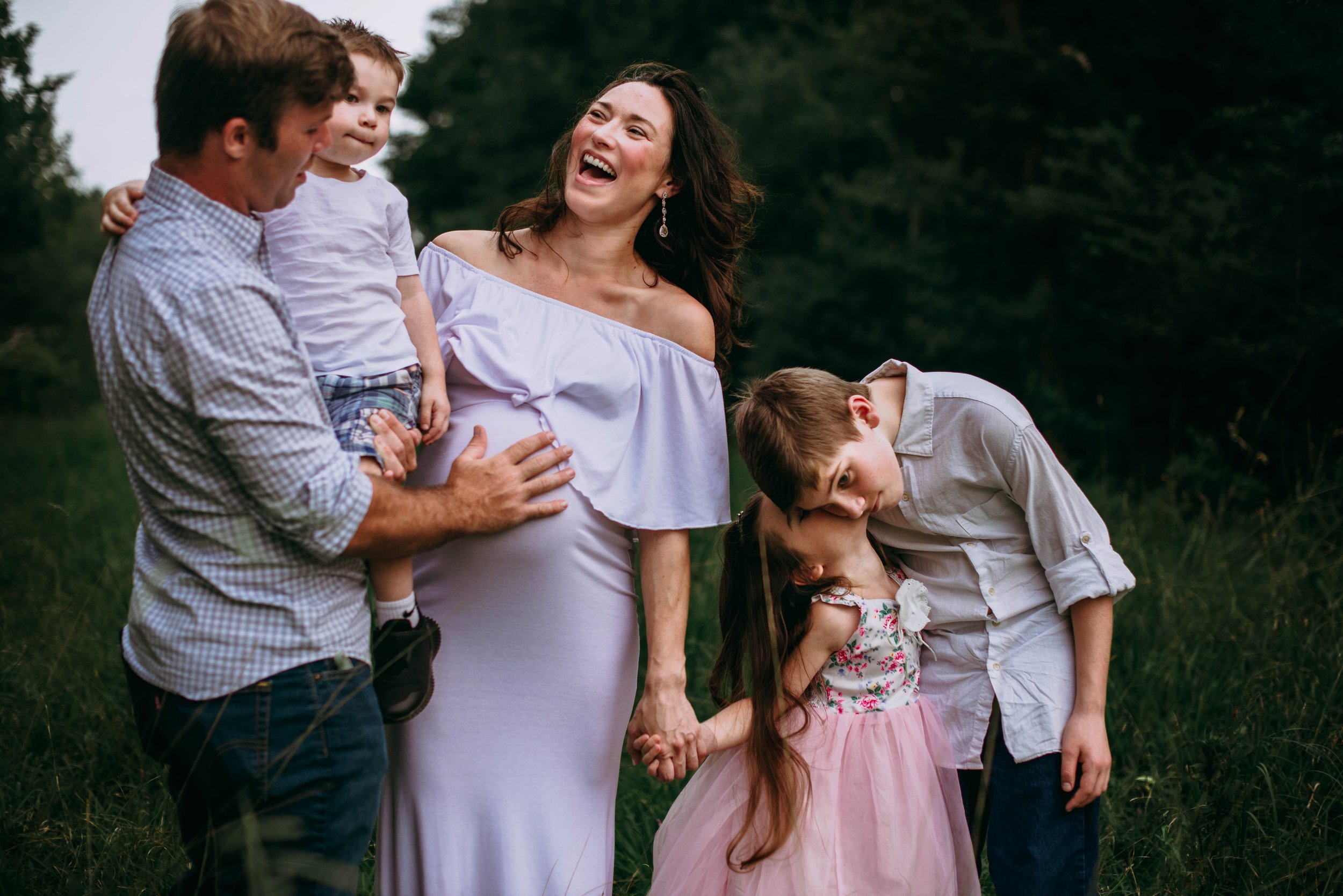 A family of five, including a woman, two boys, a girl, and a man, standing outdoors on a grassy area. The woman is laughing and holding hands with the girl, who is giving the woman a kiss on the lips. The man is holding a young boy, and an older boy 