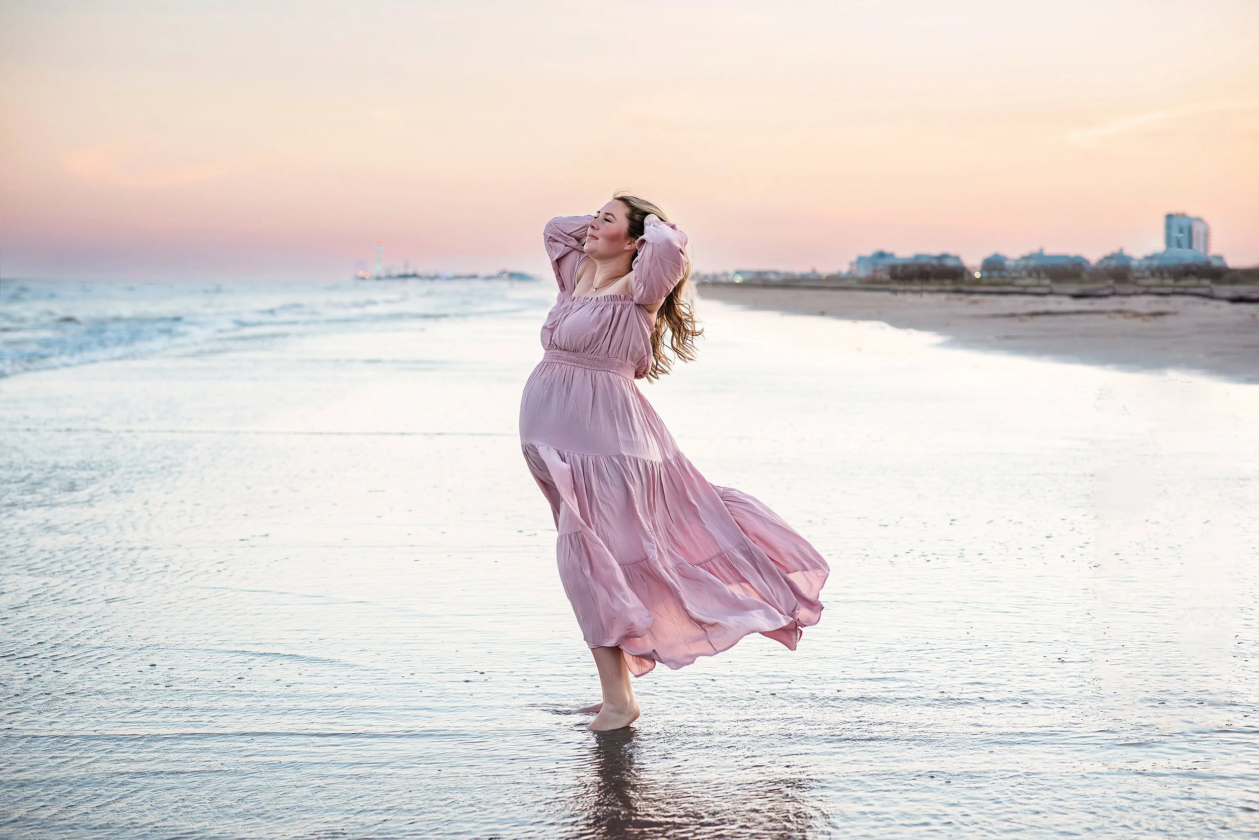 Pregnant woman in a flowing pink dress standing on the beach at sunset during a dreamy maternity photography session in Galveston, Texas by Christy White Photography.